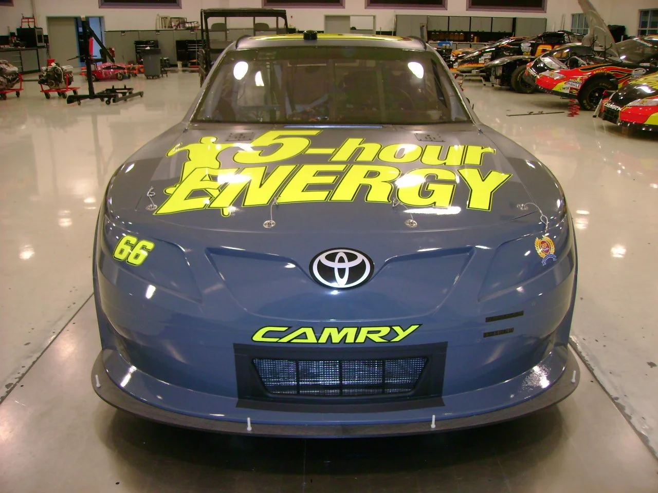 A gray Toyota Camry race car with yellow and black sponsor graphics, including the phrase '5-hour ENERGY,' the number 66, and the word 'CAMRY' on the front. The car is inside a garage or showroom with other race cars and equipment visible in the back