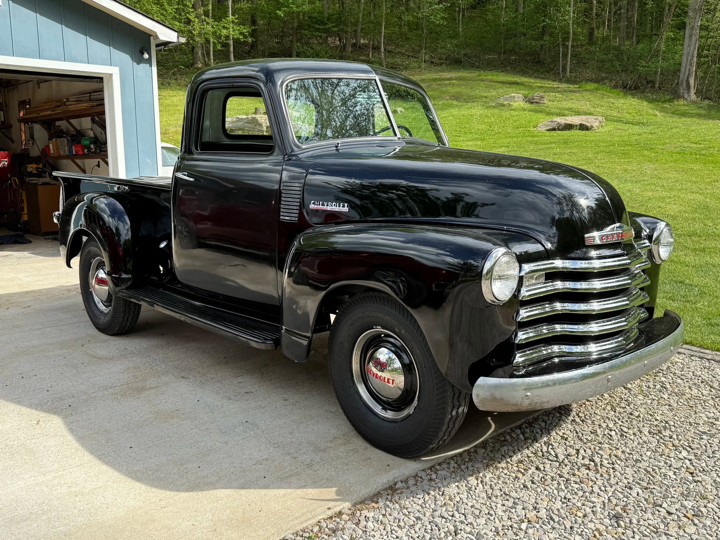 A black vintage Chevrolet pickup truck parked on a driveway next to a blue shed, with lush green trees and grass in the background.