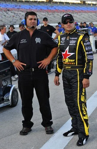 Two men, one in a racing suit and the other in casual attire with a headset, standing on a race track with racing teams and equipment in the background.