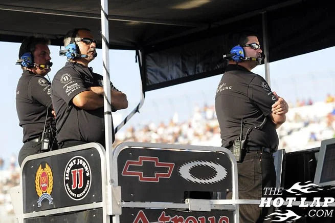Three team members standing near a race track, observing during a racing event; they are wearing black shirts, headsets, and sunglasses.