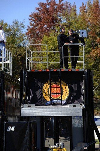 Two men standing on a raised platform with a railing, speaking into microphones at an outdoor event with trees in the background.