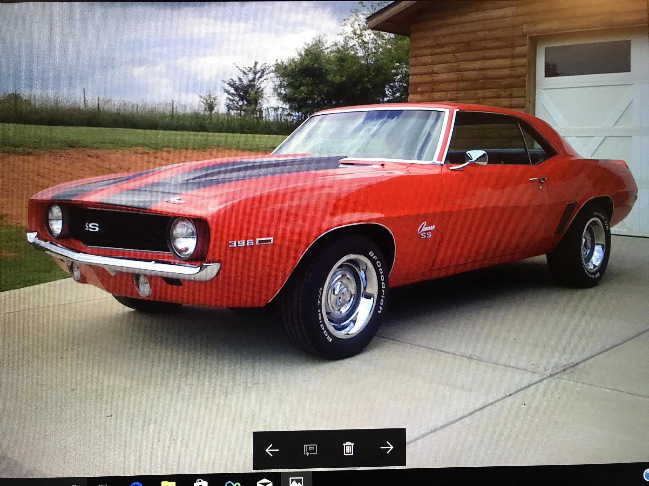 A red Chevrolet Camaro SS car with black racing stripes on the hood, parked outside a house with a garage door, next to a grassy yard and trees in the background.