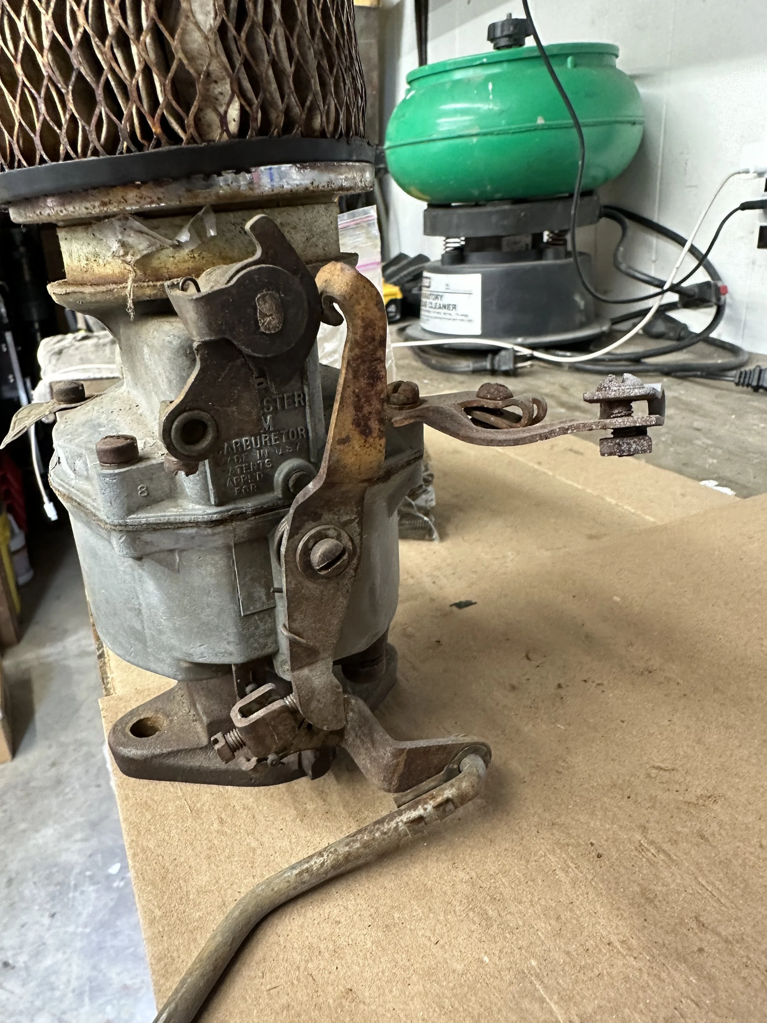 Close-up of an old mechanical carburetor with rusted parts on a workbench, with a green air filter in the background.