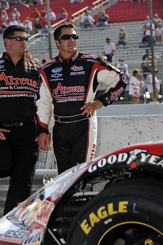 Two men in racing suits and sunglasses standing near a race car at a track, with spectators in the background.