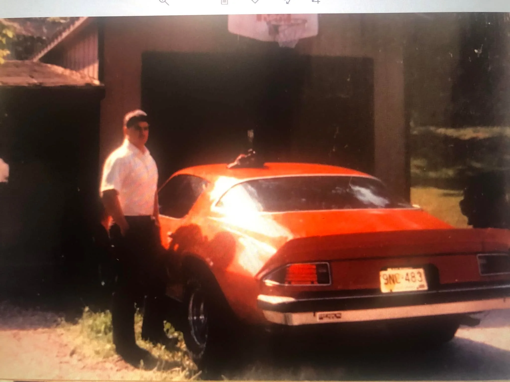 Photo of an orange car at night with a man standing nearby. The car has a Texas license plate and the scene appears to be in a residential area with grass and trees.