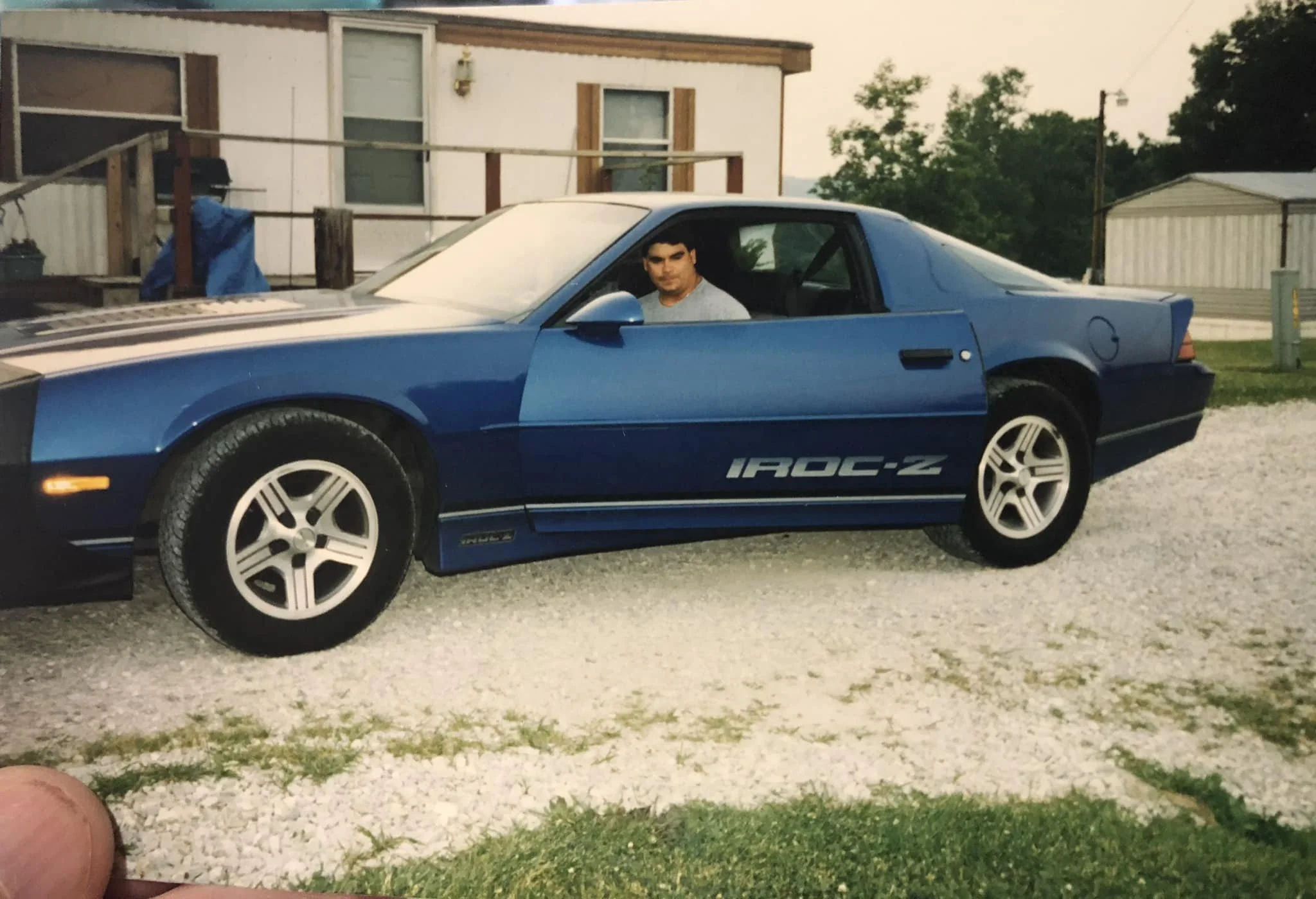 A man sitting in a blue sports car with 'IROC-Z' written on the side, parked outside a house with a porch, windows, and trees in the background.