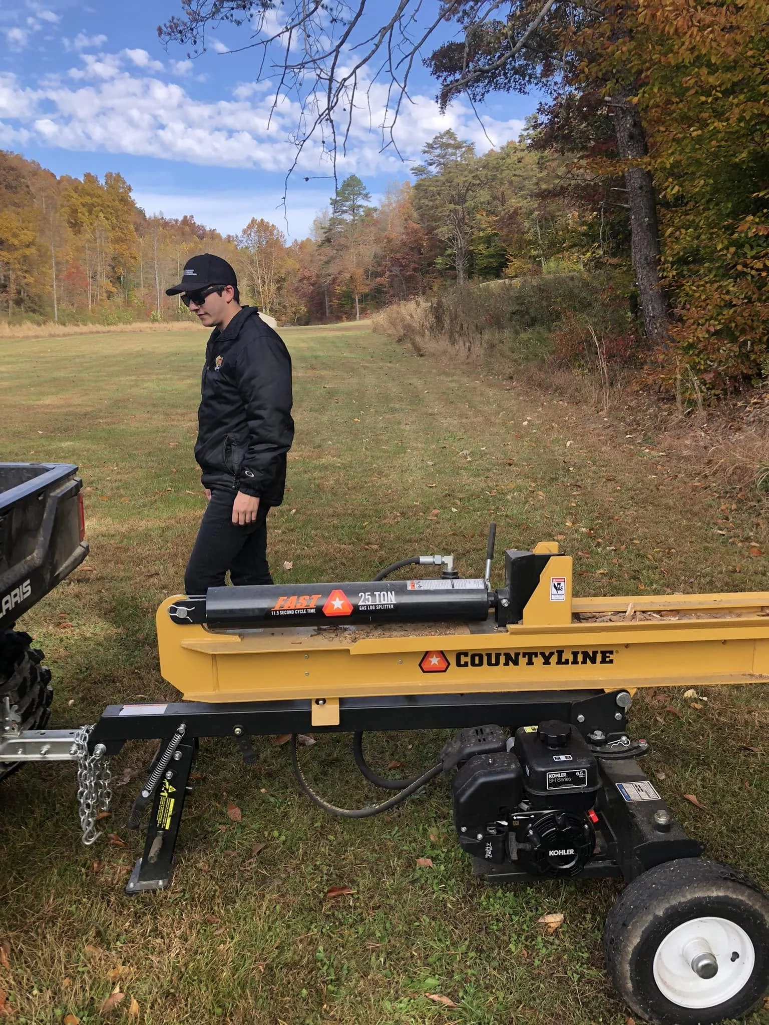 A man in a black jacket and cap standing in a grassy area, with fall trees in the background, near a yellow CountyLine log splitter and other equipment.