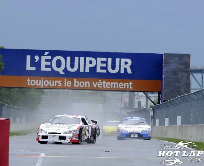 Race cars driving on a wet track with spray behind them, passing under a blue and orange sign that reads "L'ÉQUIPEUR toujours le bon vêtement."
