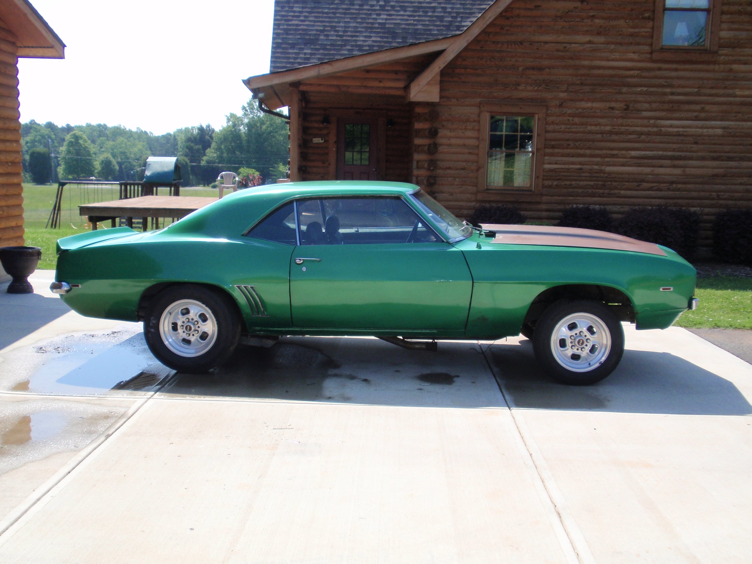 A green vintage car parked on a concrete driveway in front of a wooden house with a shingled roof and windows, with a grassy lawn and trees in the background.
