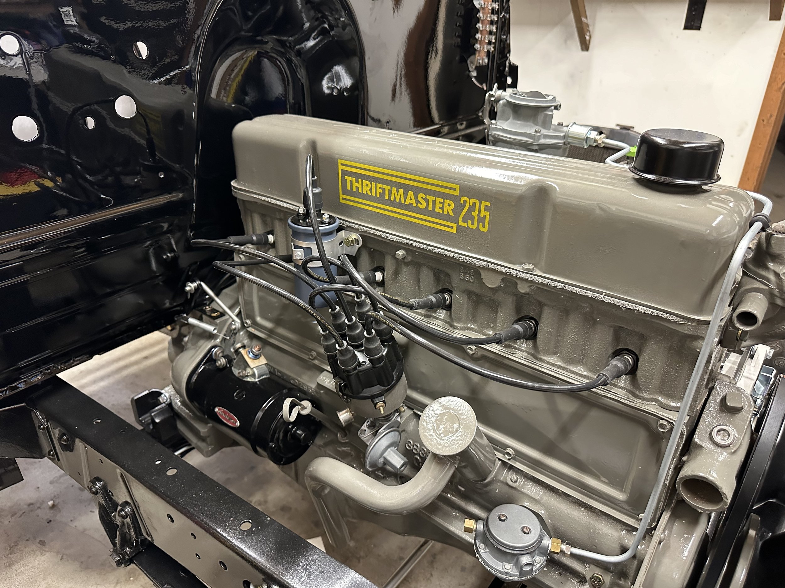 A close-up view of a vintage engine labeled 'Thriftmaster 235,' installed in a vehicle with black painted body panels.