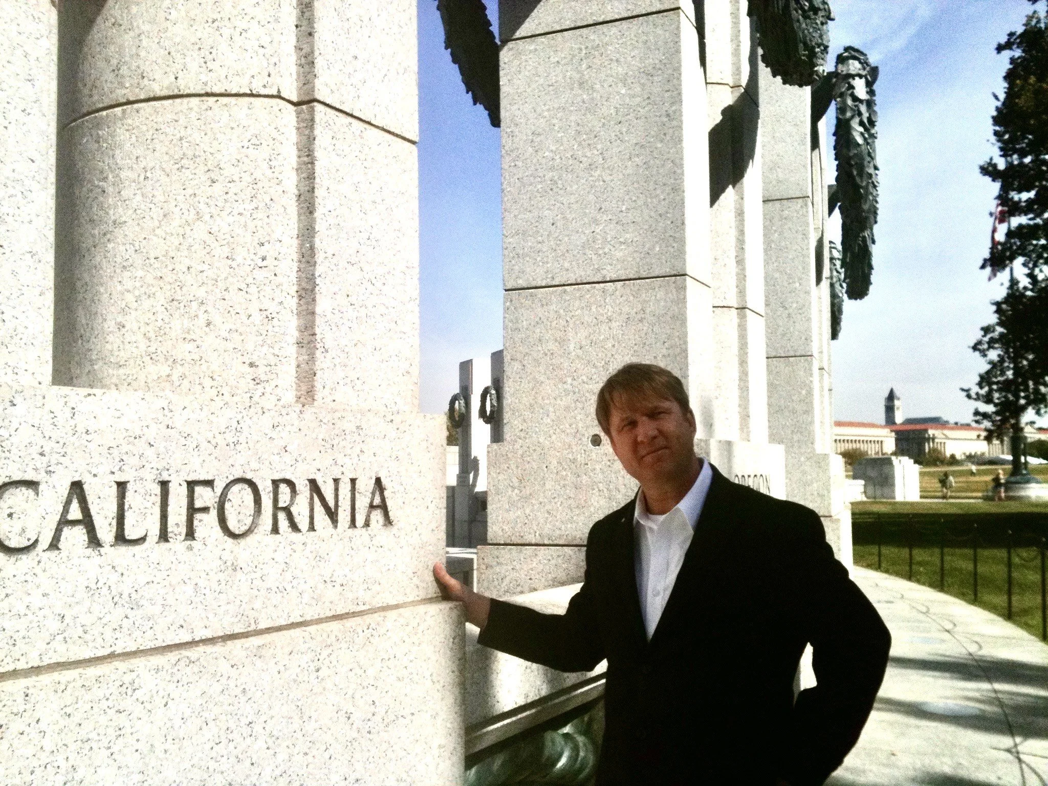 A man in a black suit and white shirt standing next to a large stone monument with the word 'California' engraved on it, in an outdoor park area.