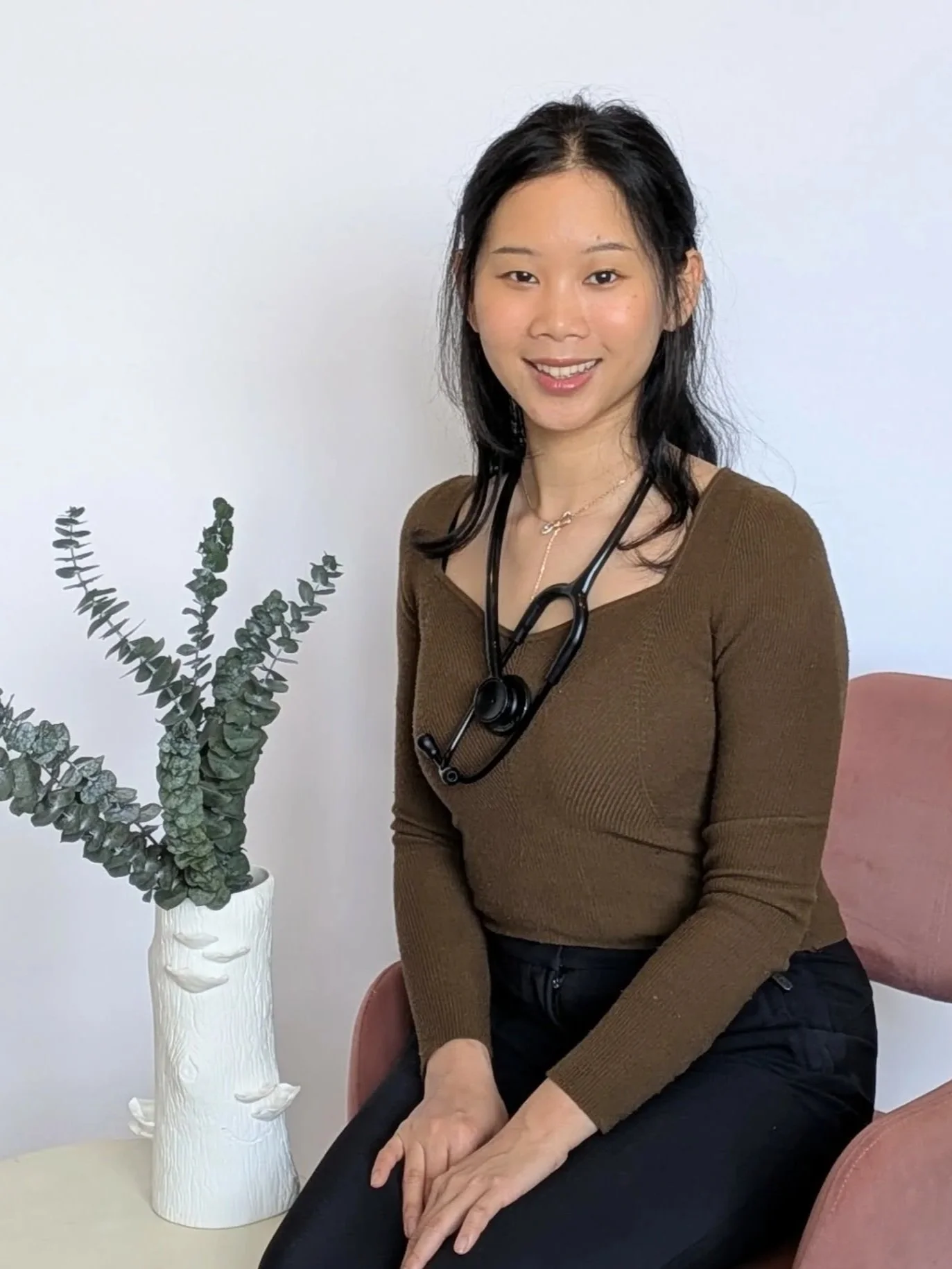 A smiling woman with black hair wearing a brown top and black pants, sitting on a pink chair beside a white vase with a dark green plant.