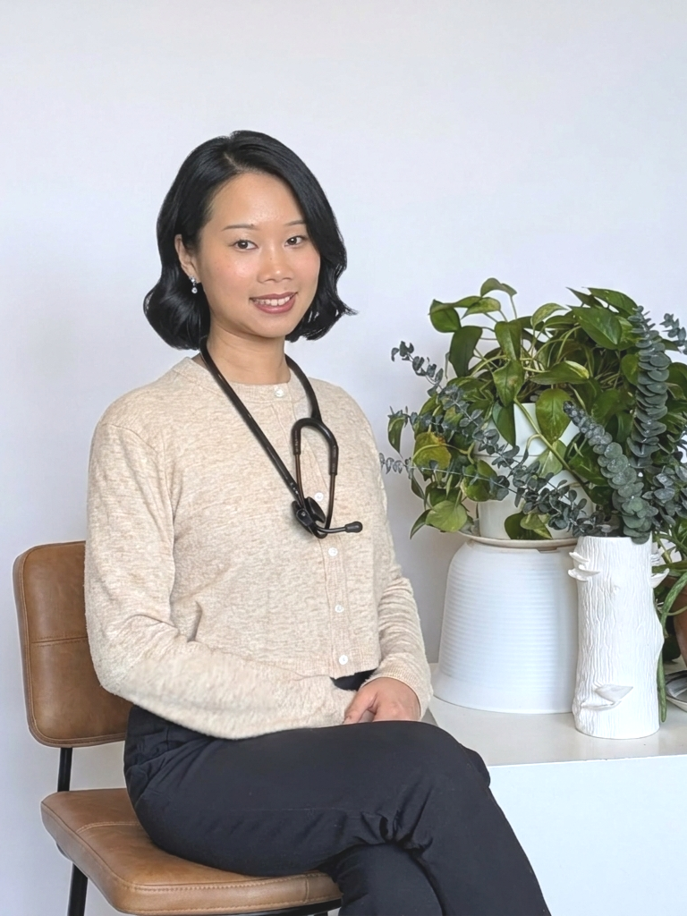 A woman with shoulder-length black hair, wearing a beige sweater and black pants, sitting on a brown chair next to a white table with large potted green plants.