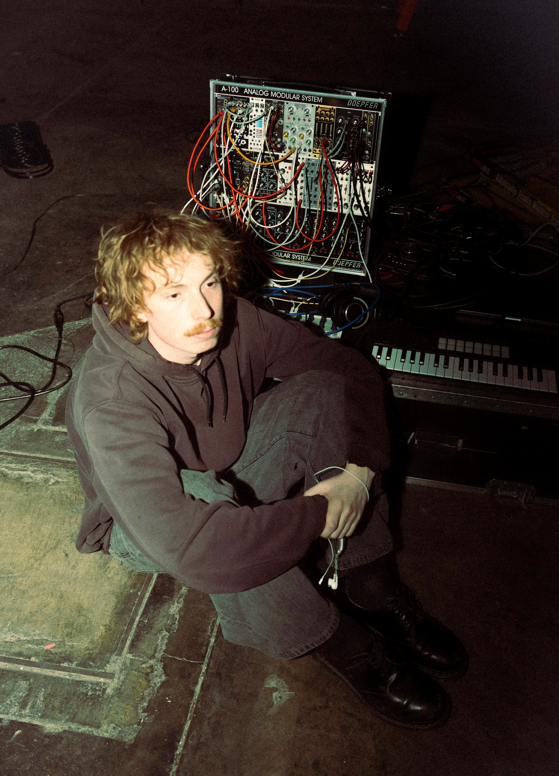A man with curly hair and a mustache, wearing a hoodie, sits on the floor next to a keyboard and modular synthesizer with numerous cables.