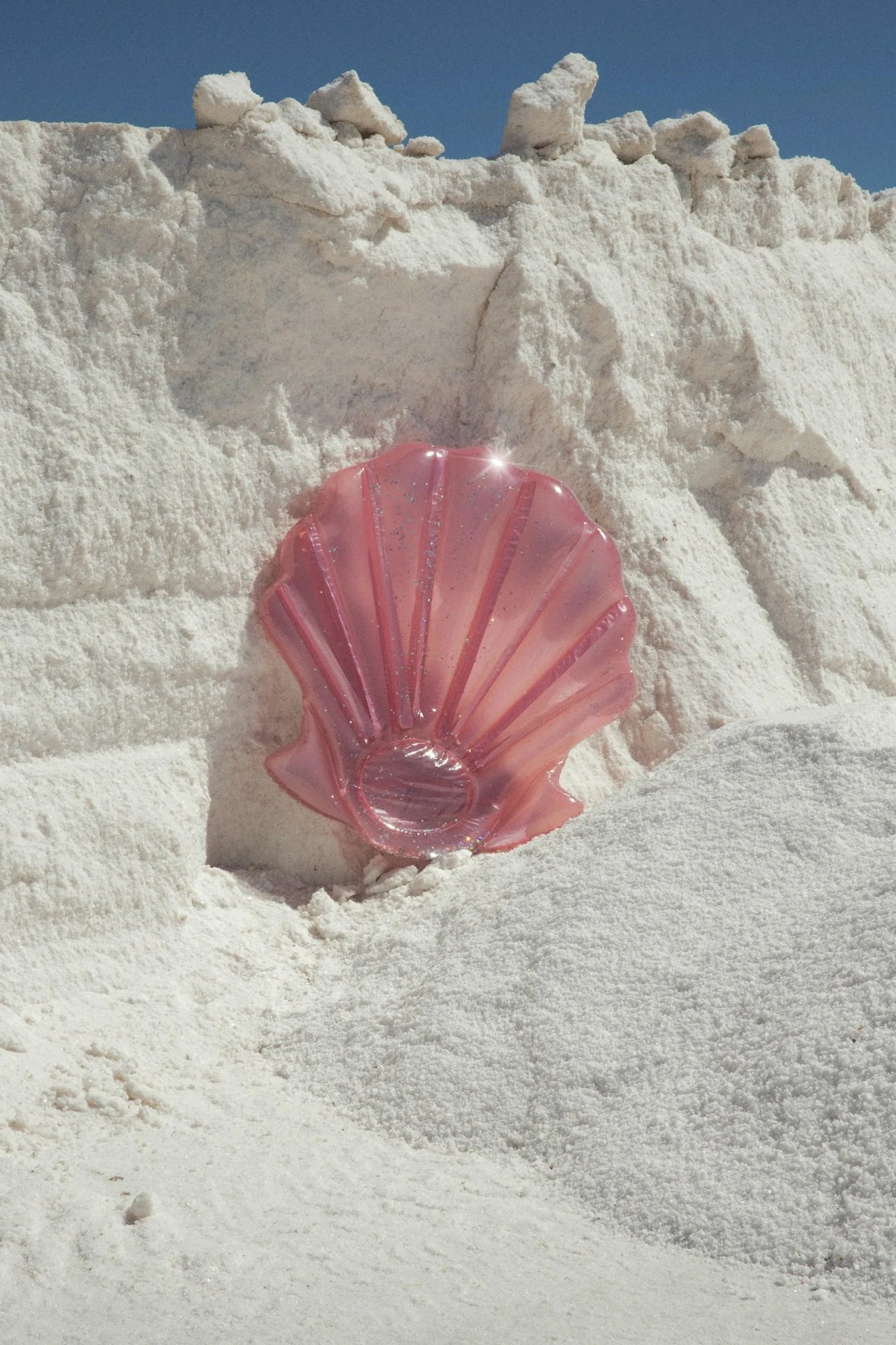 Pink seashell-shaped glass sculpture in white sand under a blue sky