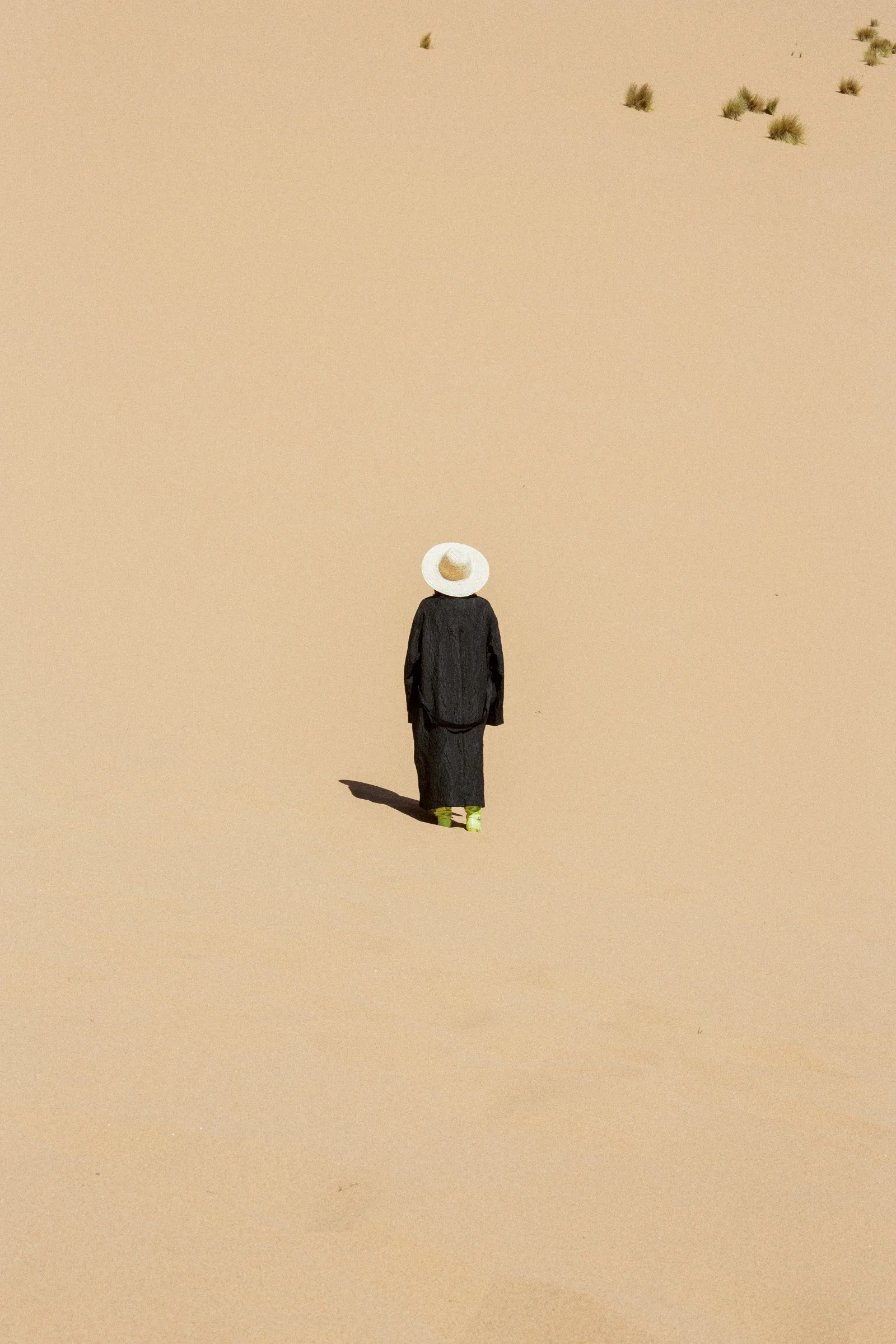 A person wearing a white hat, dark coat, and green shoes walking in a sandy desert landscape with sparse desert plants.