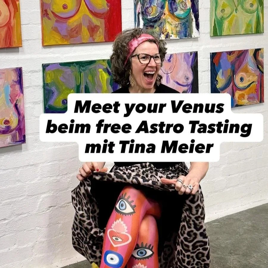  Astrologin Tina sitzt vor einer Wand aus bunten Brust-Portraits, selbst in knalligen Farben angezogen und hebt ihren Leorock an. Ihre Beine stecken in Strumpfhosen mit Augen drauf 