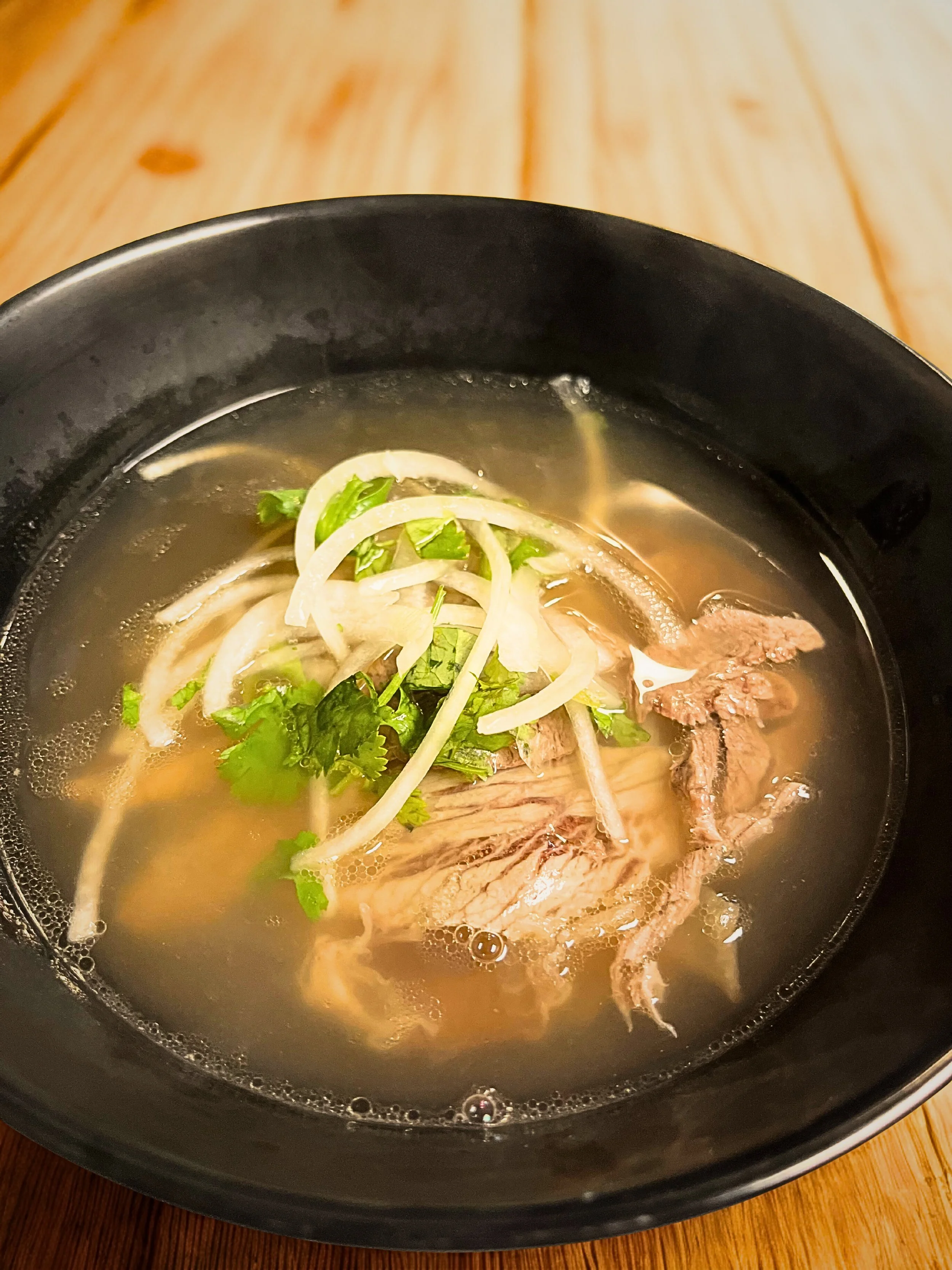 A bowl of hot ramen soup with sliced beef, onions, green herbs, and noodles in a clear broth on a wooden table.