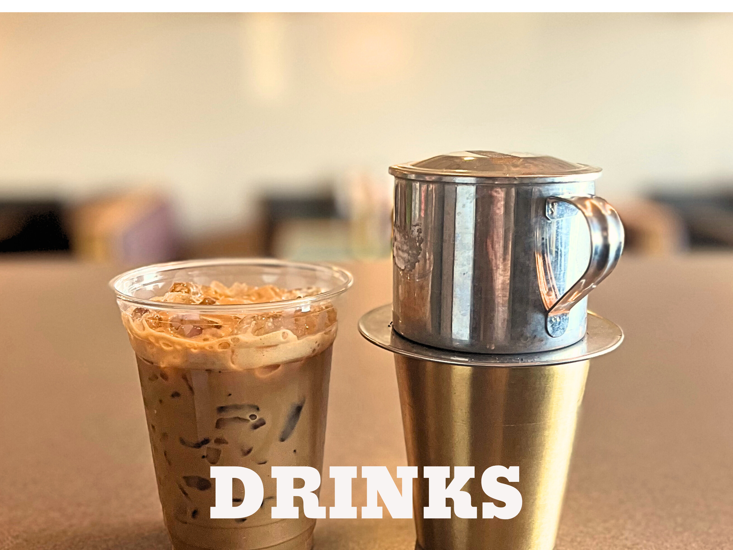 A plastic cup filled with iced coffee and a metal coffee mug on a gold-colored tumbler, with the word 'DRINKS' in bold white letters at the bottom.