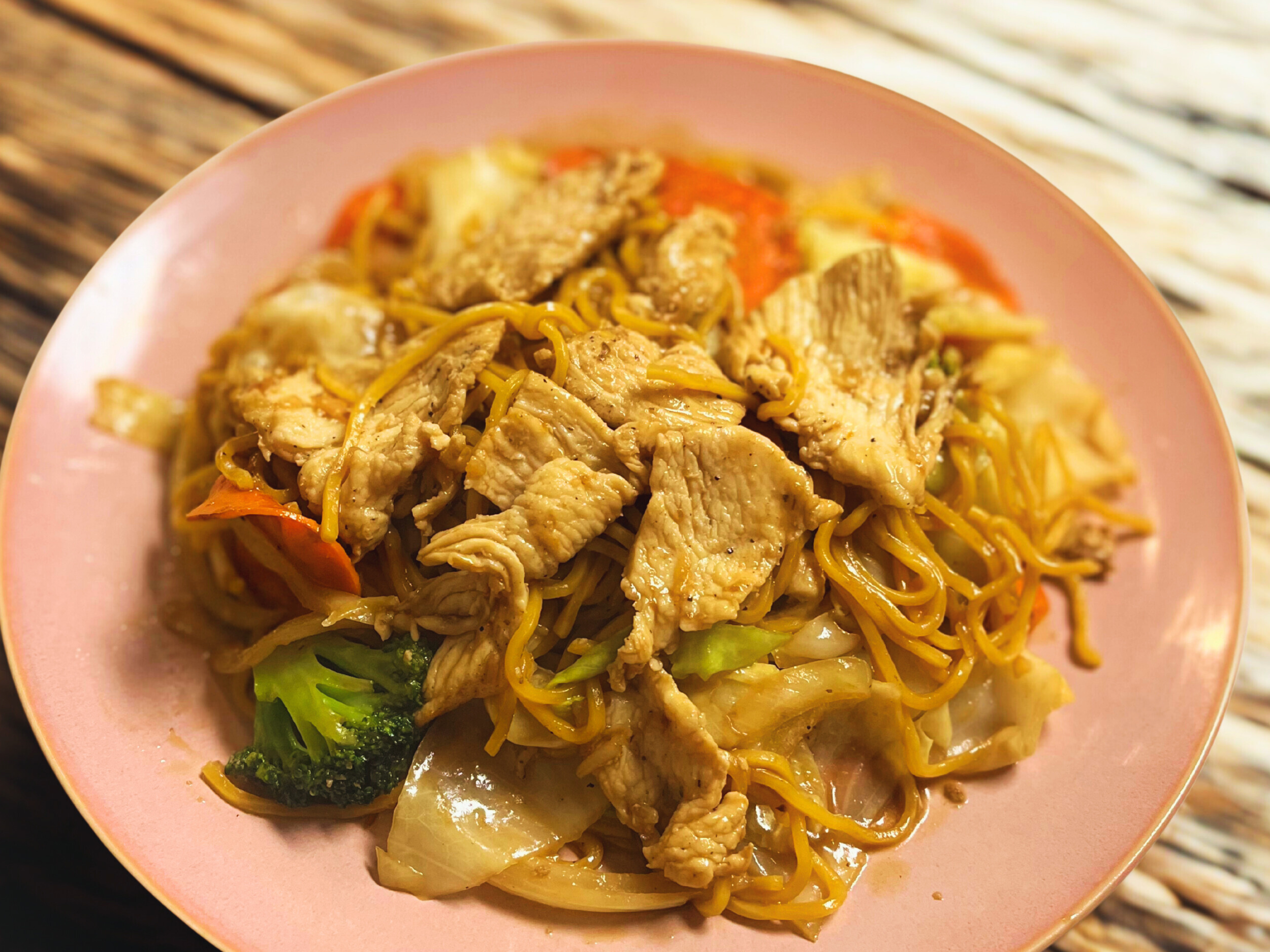 A pink bowl filled with stir-fried noodles, sliced chicken, broccoli, and vegetables on a woven bamboo surface.