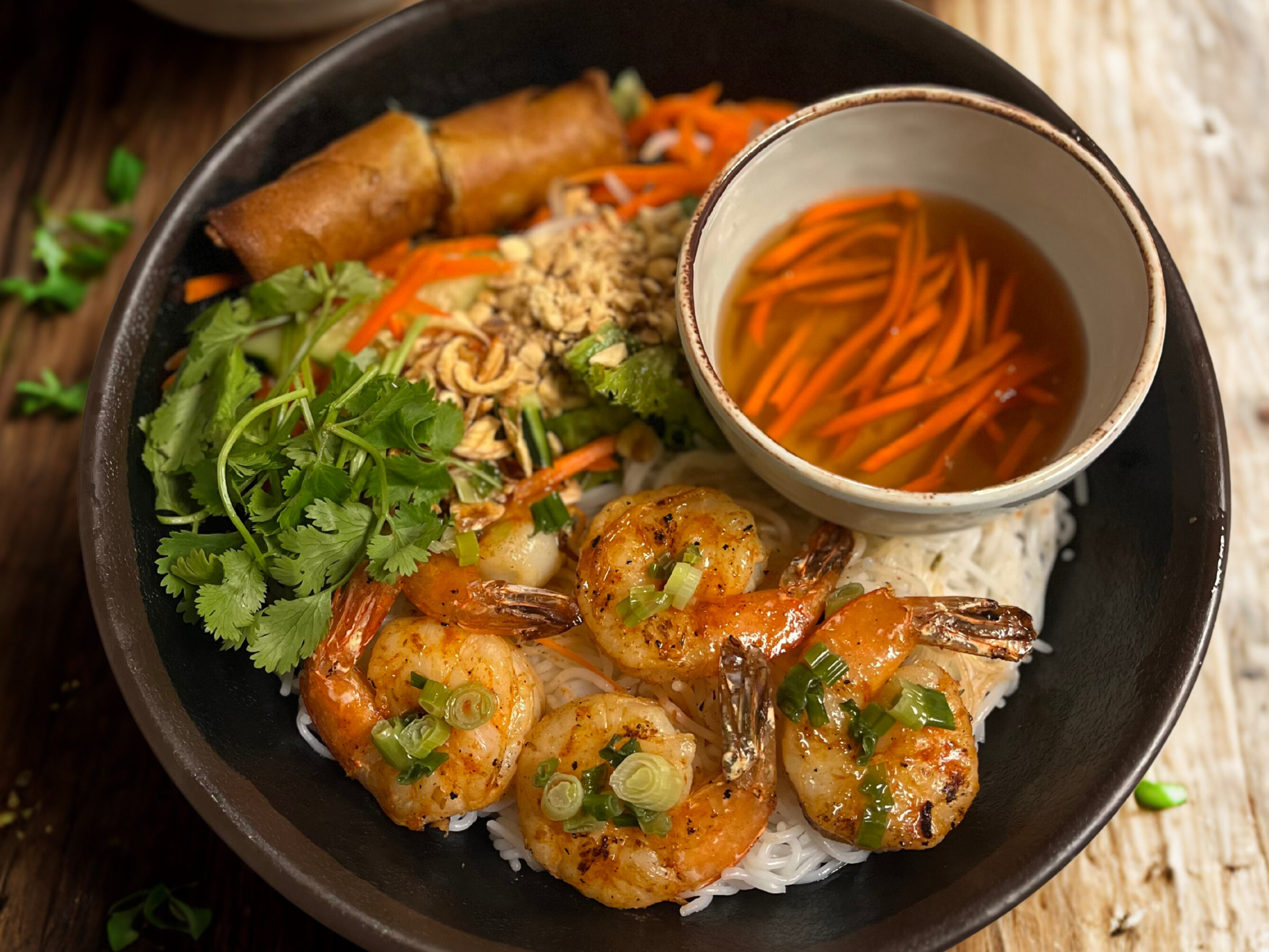 Bowl of Vietnamese shrimp spring rolls with herbs, rice noodles, and vegetables, served with a side cup of dipping sauce containing shredded carrots.