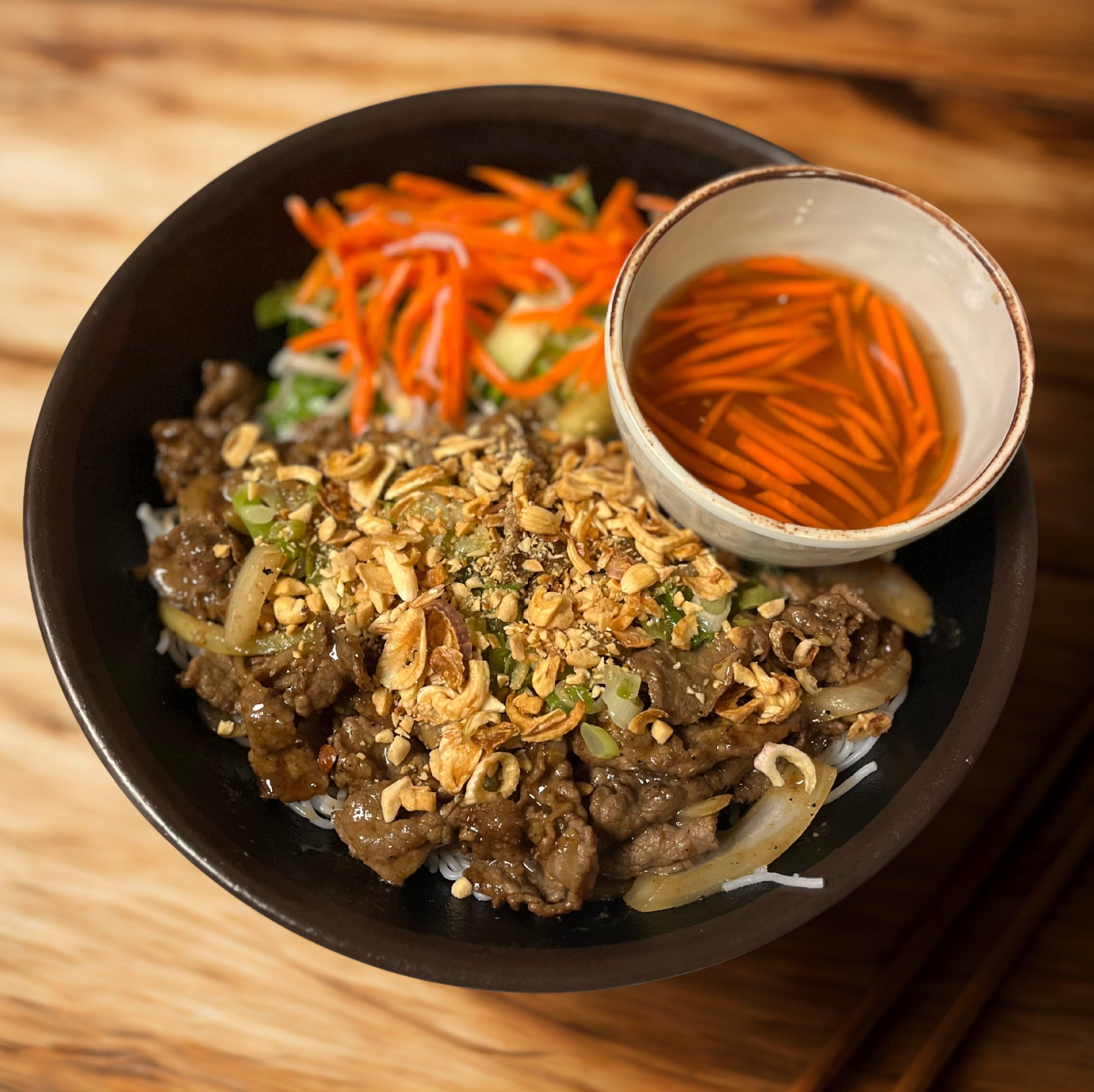Bowl of Asian beef stir-fry with chopped nuts, vegetables, shredded carrots, and a side of shredded carrots and lettuce, with a small bowl of soy sauce on top, all served on a wooden table.