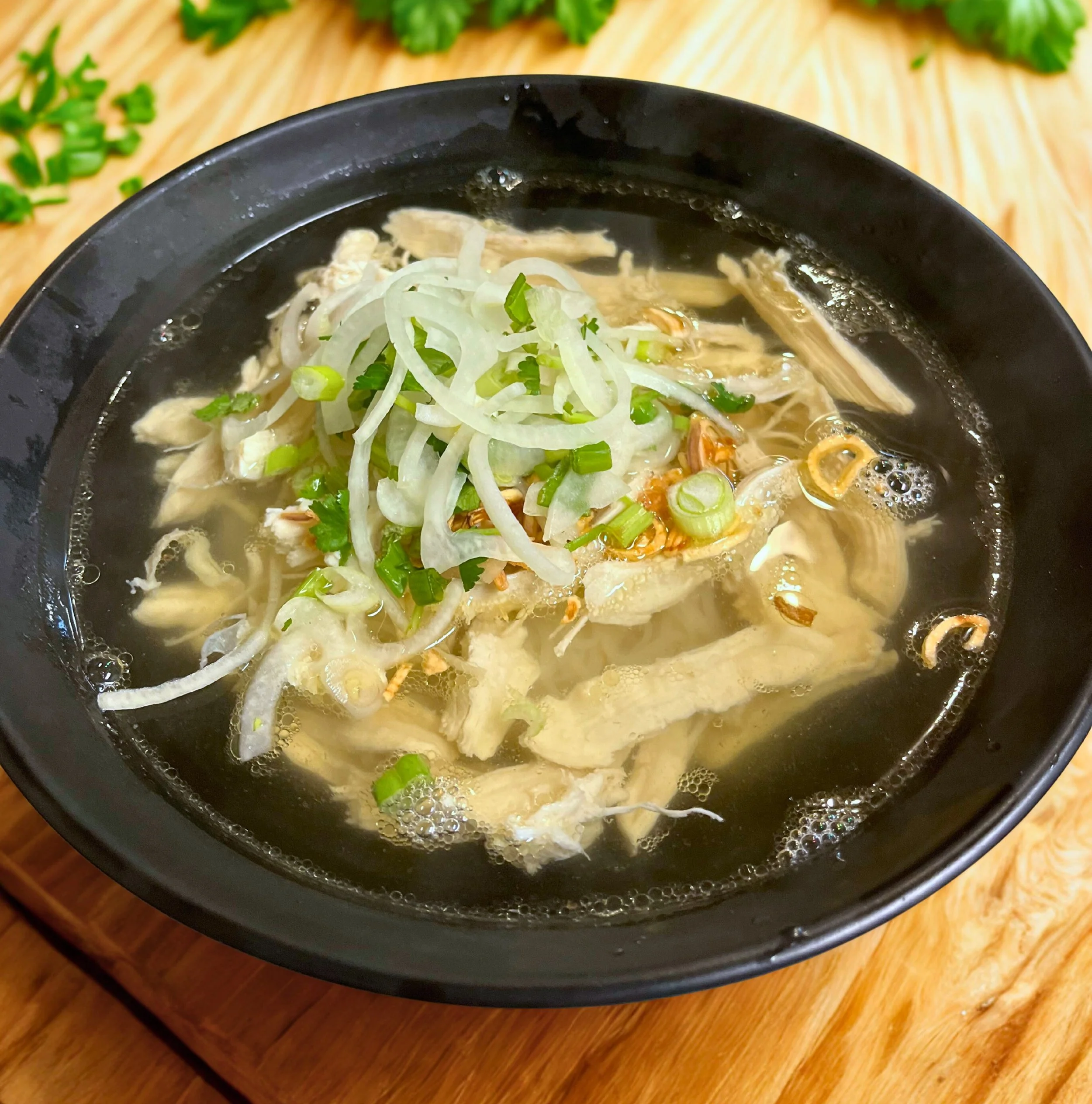 A bowl of chicken soup topped with sliced onions and chopped green onions on a wooden table.