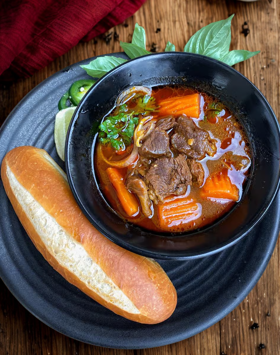 Beef stew with carrots, onions, and cilantro in a black bowl, accompanied by a bread roll on a black plate, garnished with fresh herbs and lemon slices on a wooden table.