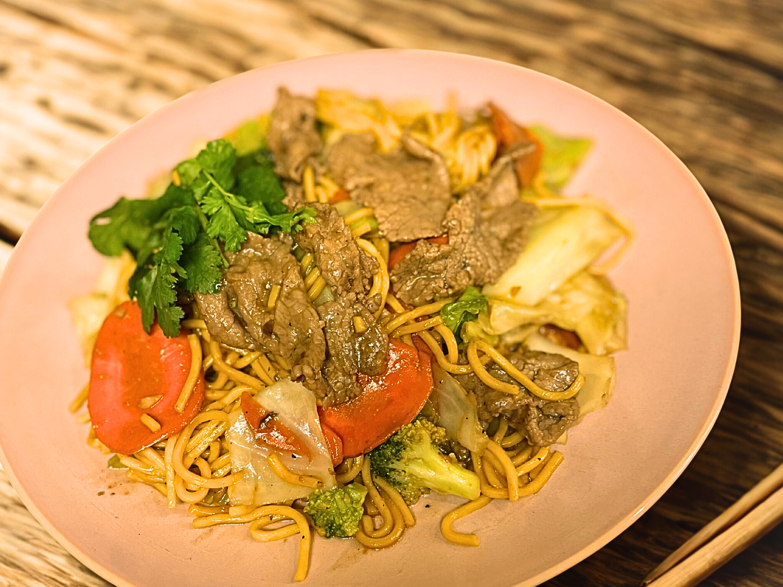 A plate of stir-fried noodles with beef, vegetables, and cilantro on a pink plate.