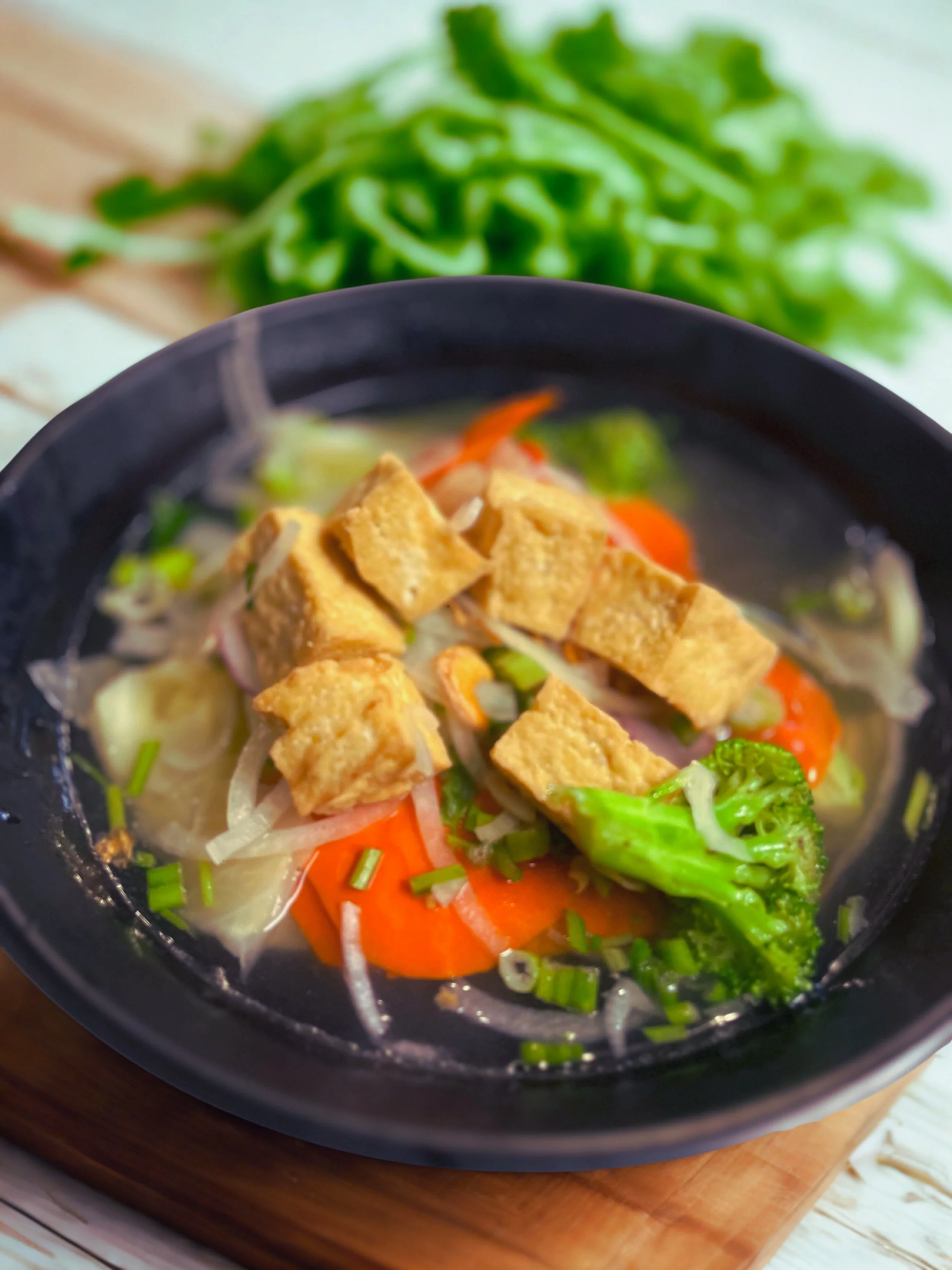 Bowl of vegetable soup with tofu, carrots, broccoli, and onions, garnished with chopped green onions, on a wooden surface with leafy greens in the background.