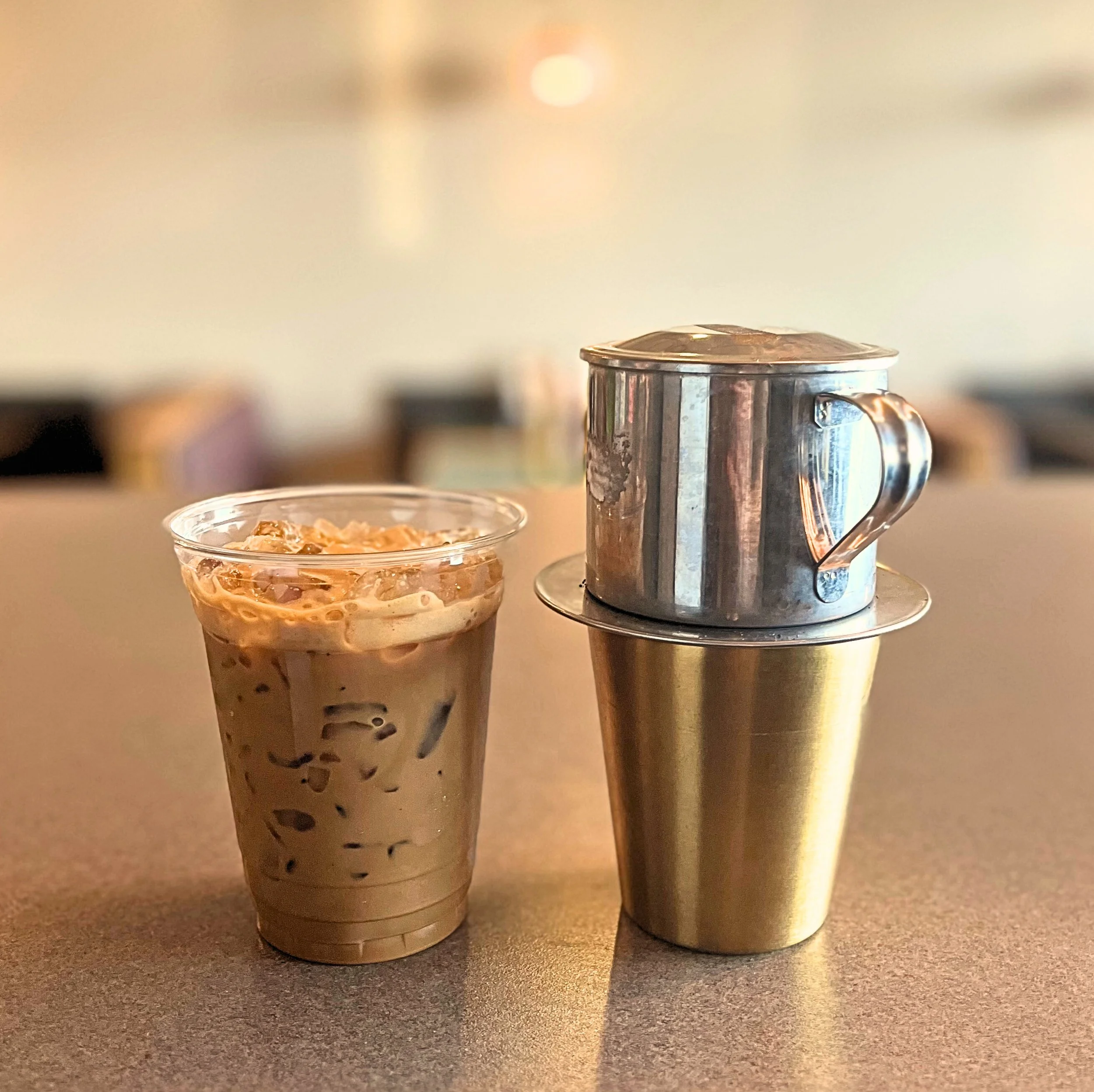 An iced coffee in a clear plastic cup next to a stainless steel coffee dripper on a metallic travel mug