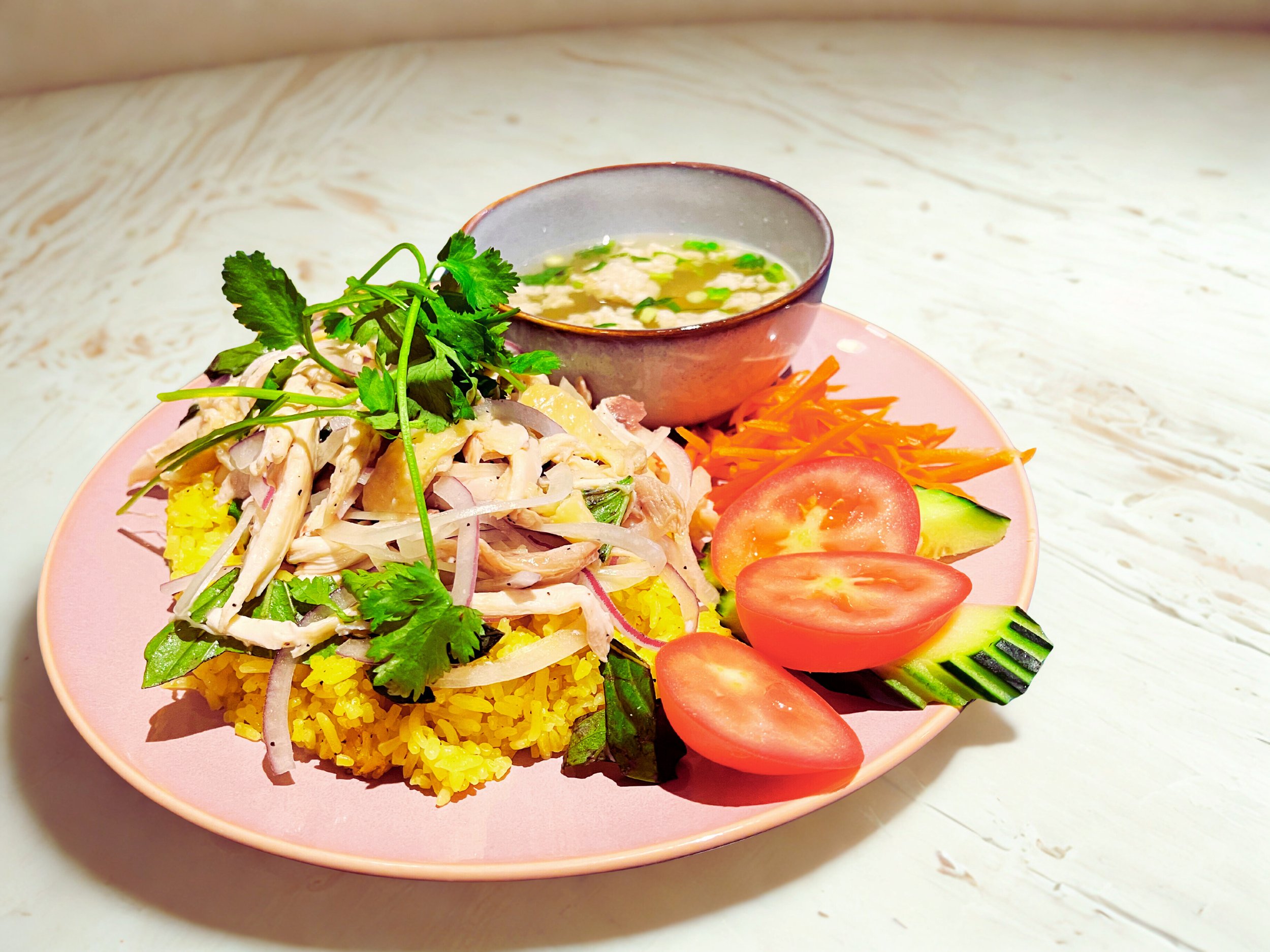 A plate of lemon rice topped with shredded chicken and herbs, served with fresh tomato, cucumber slices, and shredded carrots, accompanied by a bowl of clear soup with herbs.