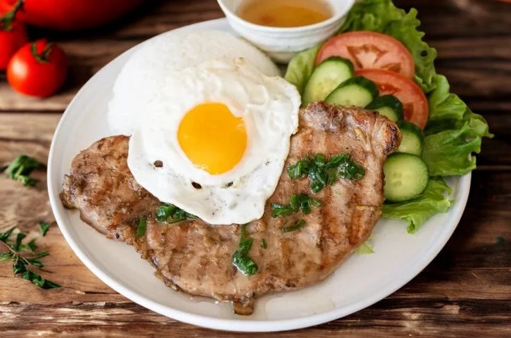 Grilled pork chop with a fried egg on top, served with sliced cucumber, tomato, lettuce, and a small bowl of dipping sauce on a white plate.