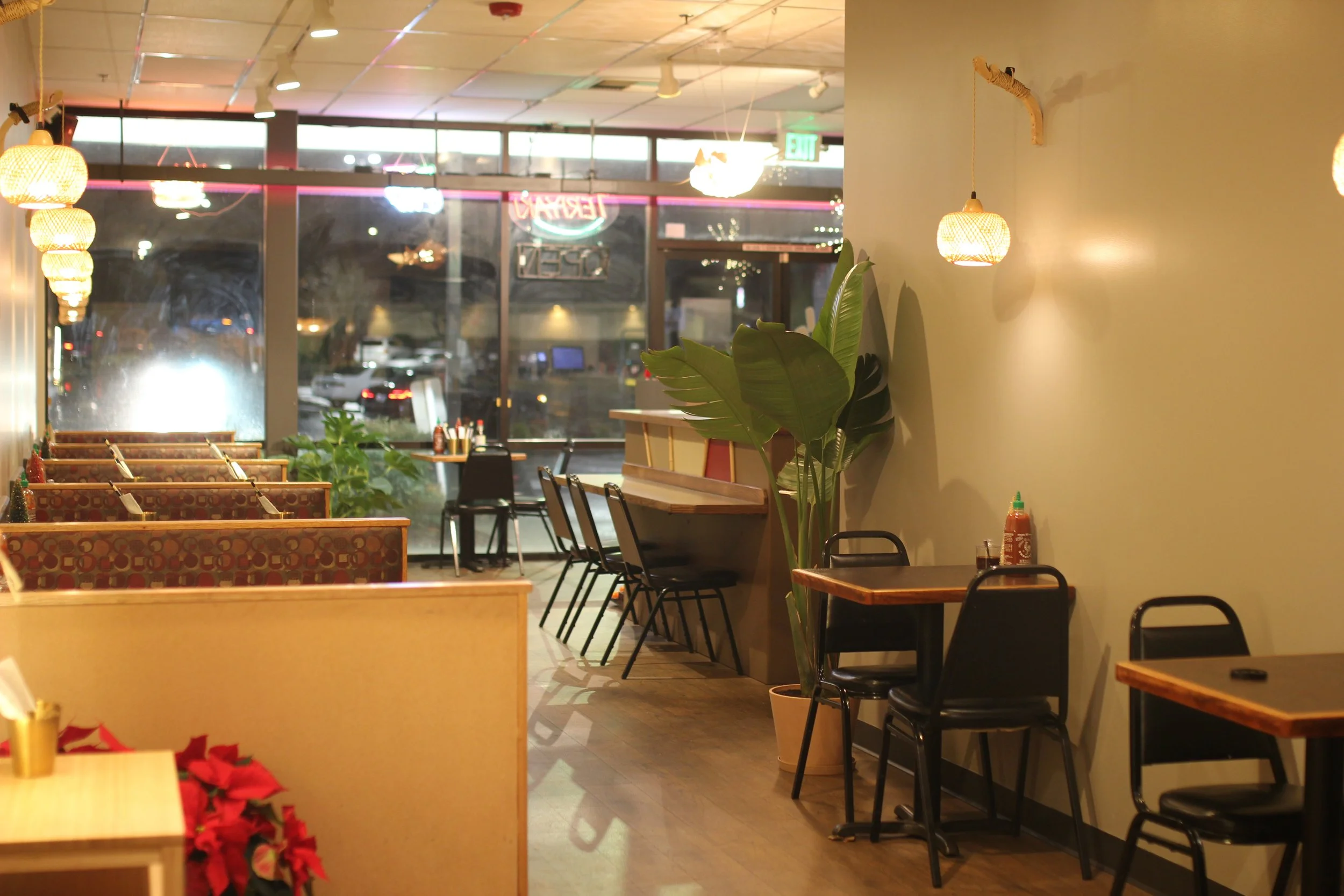 Interior of a cozy restaurant with wooden tables and black chairs, potted plants along the wall, warm lighting, and large front windows showing a parking lot outside at night.