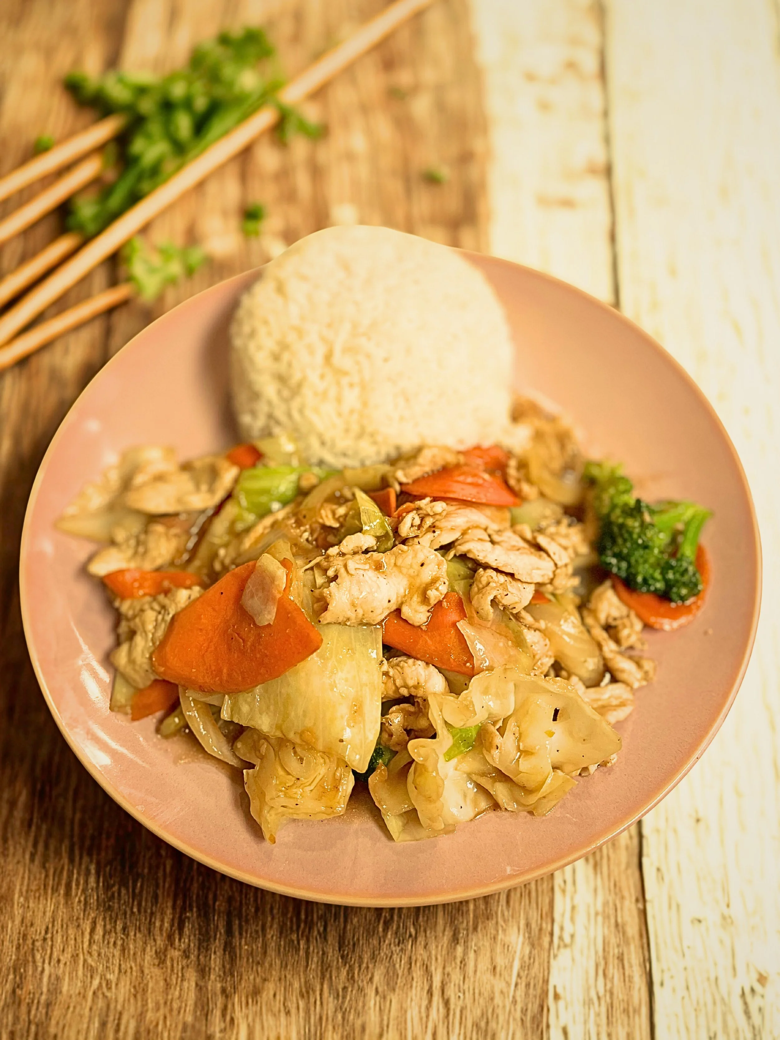 A pink bowl filled with stir-fried vegetables and sliced chicken, accompanied by a scoop of rice, on a wooden table with chopsticks and a sprig of parsley in the background.