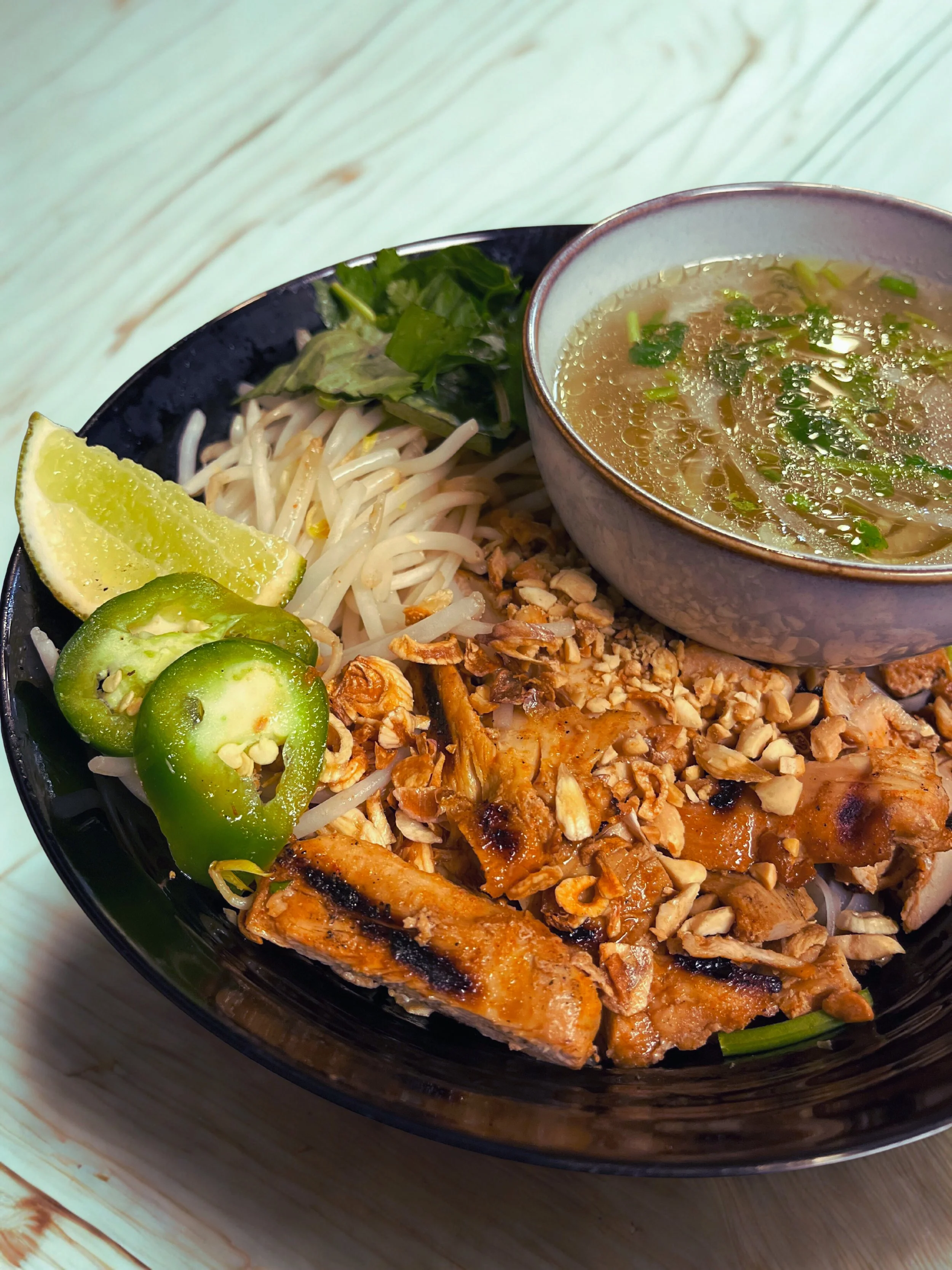 Bowl of Vietnamese Pho with grilled chicken, bean sprouts, fresh herbs, lime wedge, sliced jalapenos, and a side of broth.