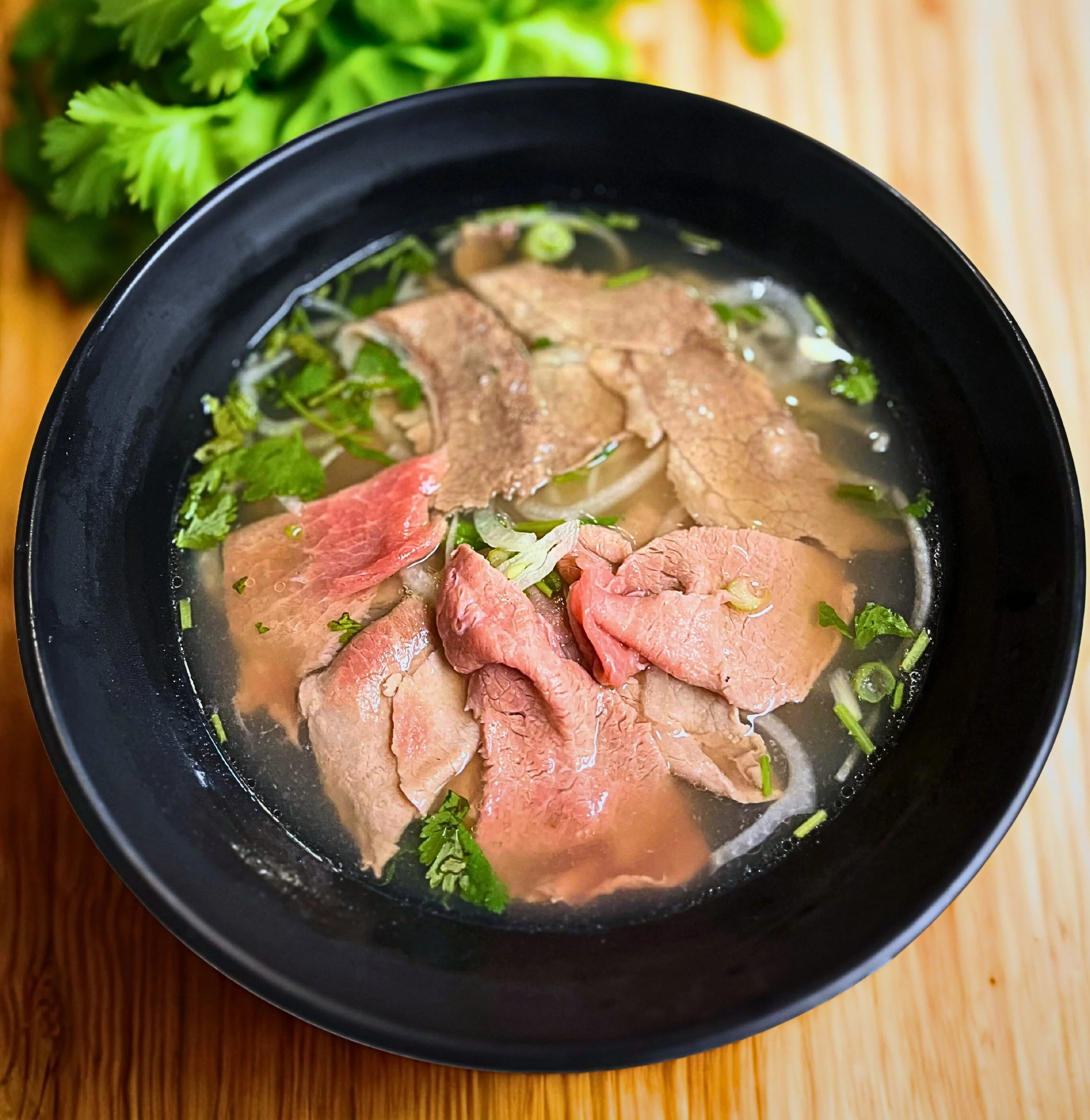 A bowl of Vietnamese pho with sliced beef, fresh herbs, and green onions, served on a wooden table.