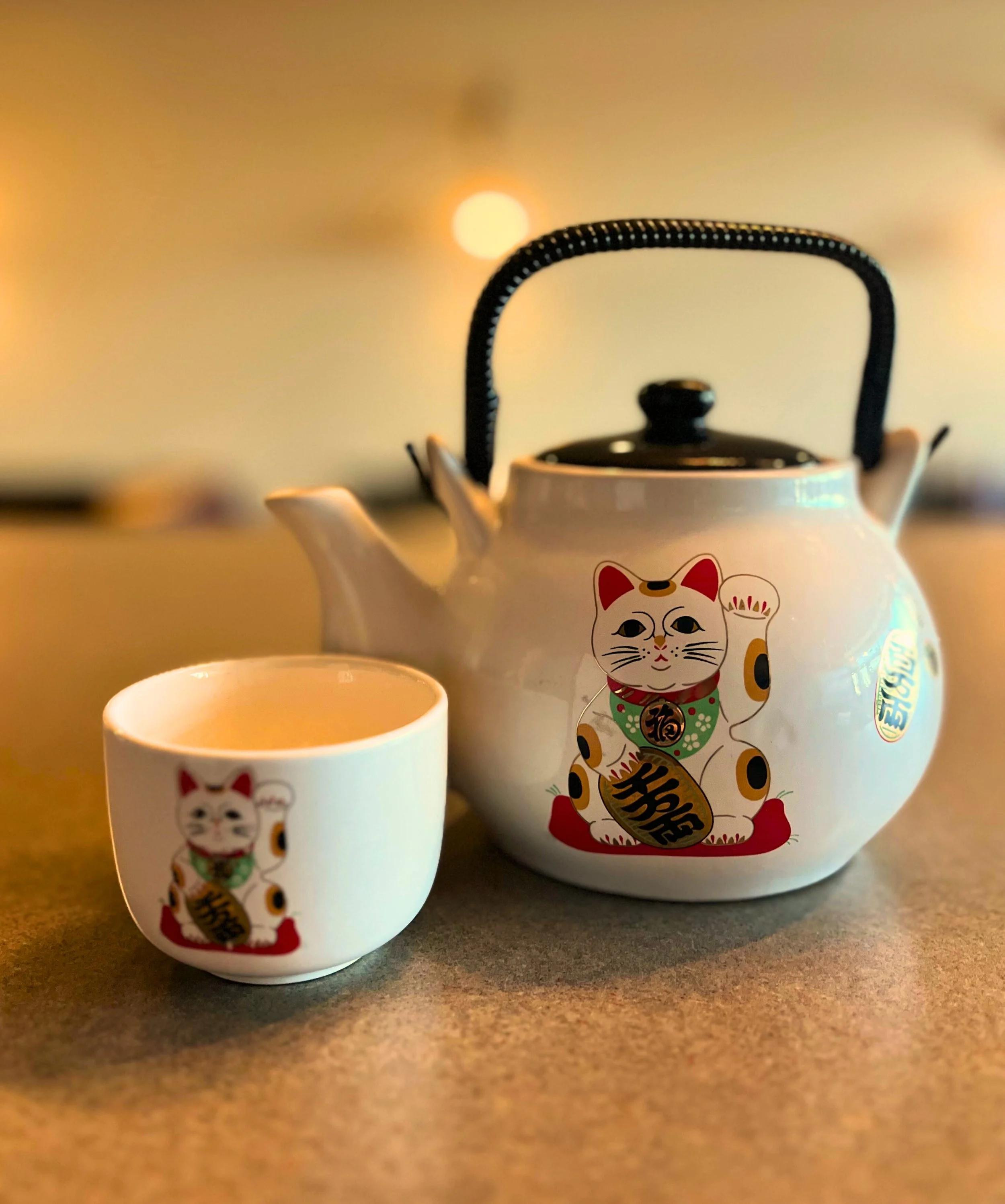 A white teapot and teacup, both decorated with a Maneki-neko (lucky cat) design, on a gray surface with blurred warm background lights.
