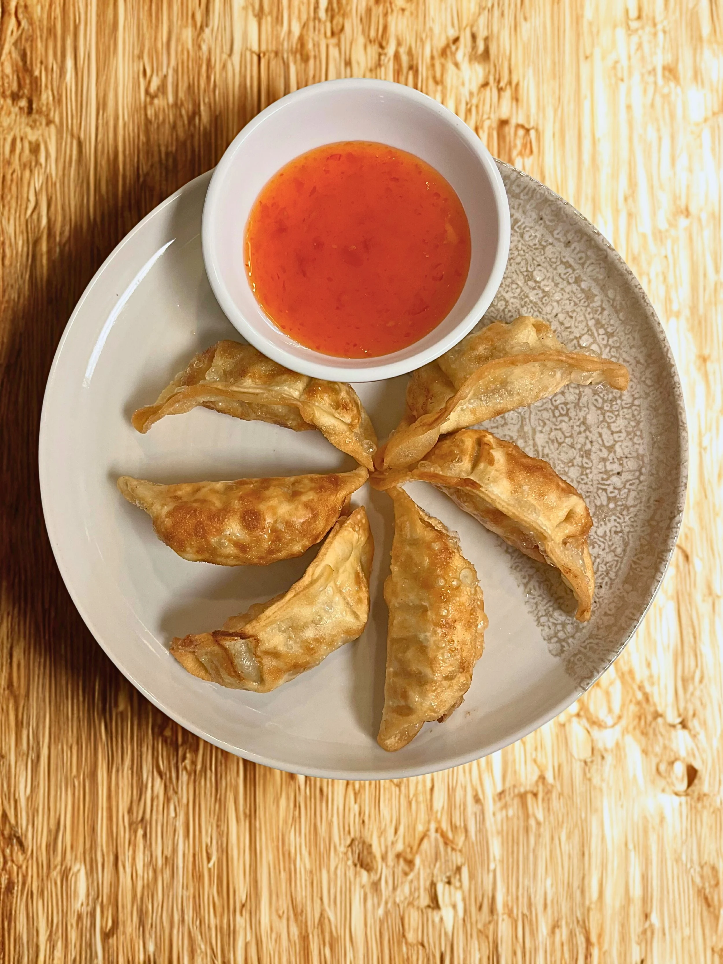 Six fried dumplings arranged in a circle on a white and textured ceramic plate, with a small bowl of orange dipping sauce.