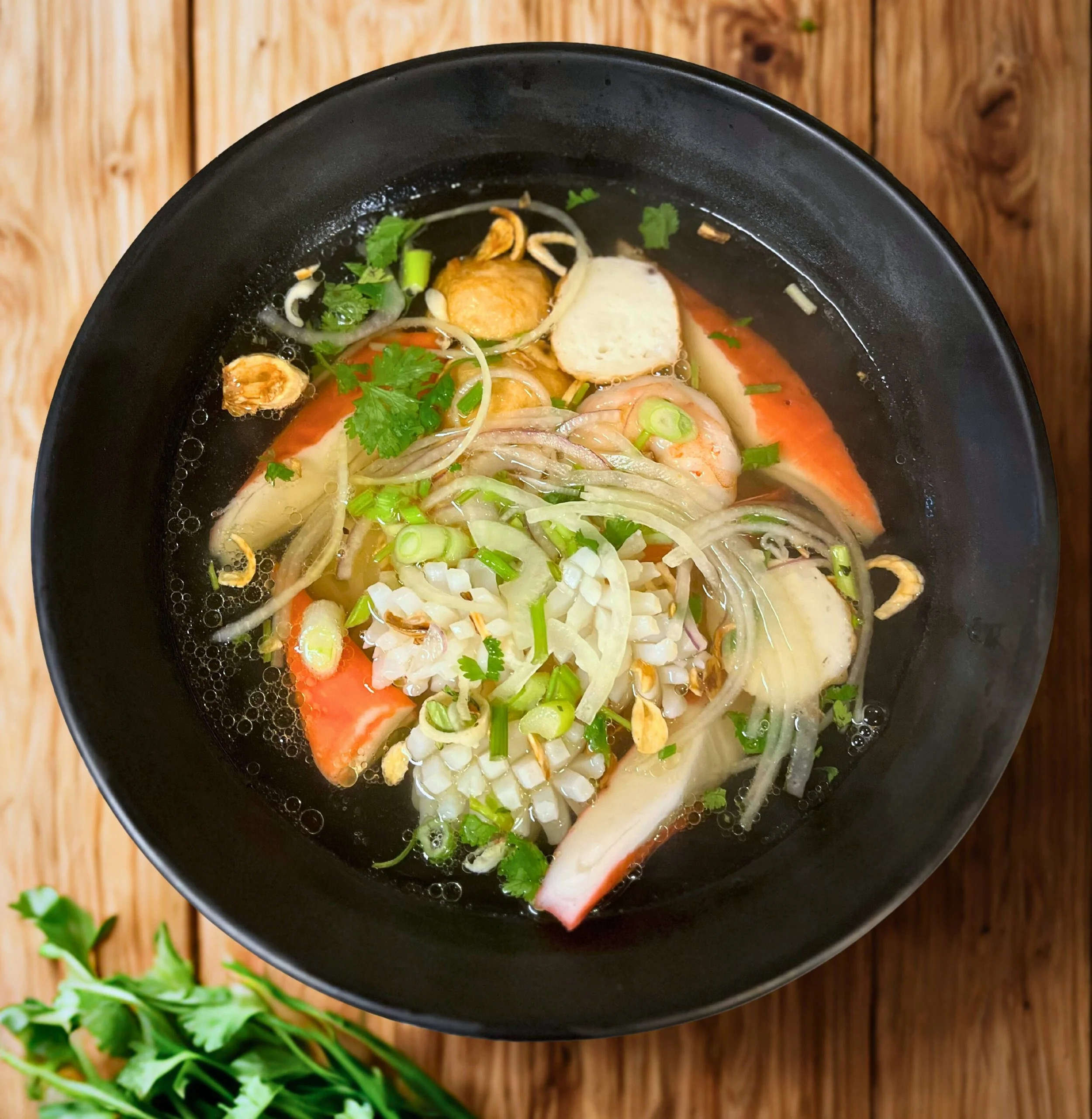 Bowl of Vietnamese noodle soup topped with cilantro, green onions, sliced onions, prawns, crab sticks, and fried garlic, served in a black bowl on a wooden table.
