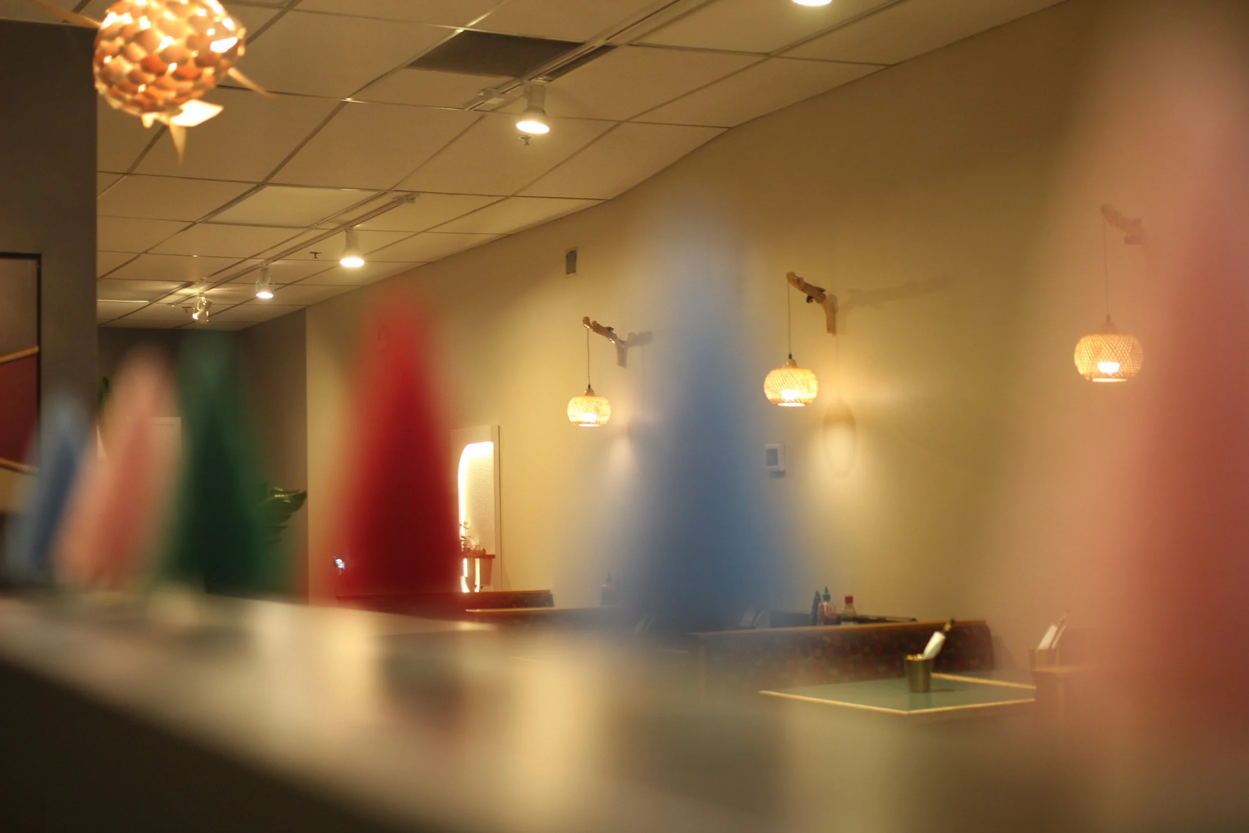 A dimly lit interior of a restaurant or café with hanging pendant lights on the walls, a blurred foreground with colorful objects, possibly napkin holders, and a few tables and chairs in the background.