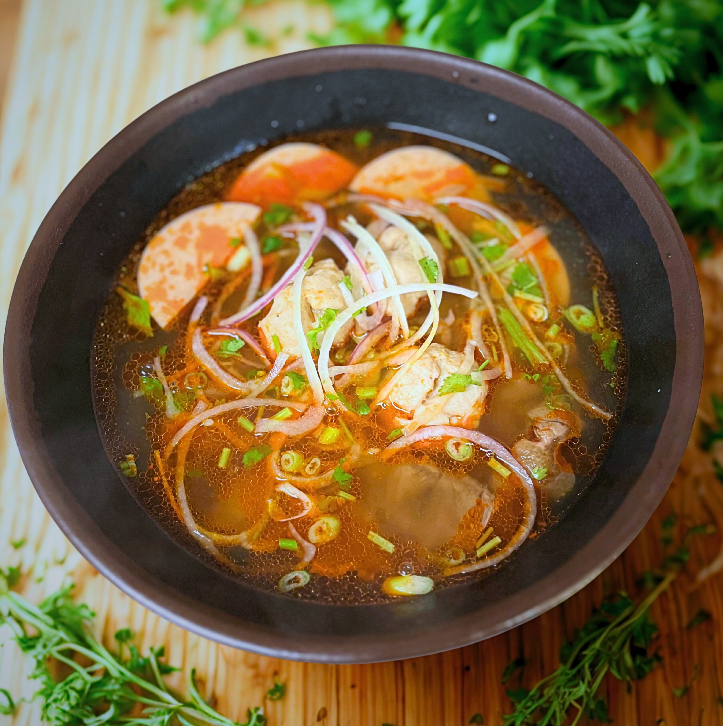 A bowl of Vietnamese Pho soup with slices of beef, boiled eggs, onions, green onions, and herbs in a dark ceramic bowl, on a wooden surface with fresh herbs around.