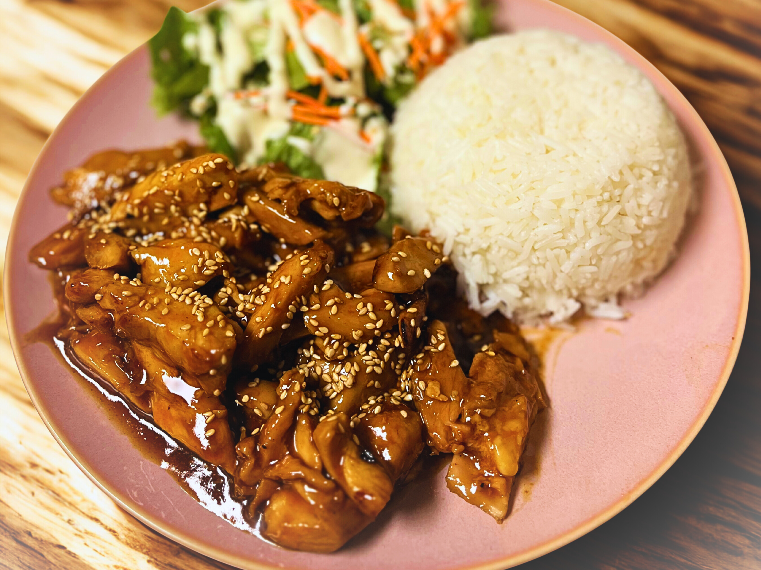 A plate of Asian-style stir-fried chicken with sesame seeds, a serving of white rice, and a side of salad with lettuce and shredded vegetables.