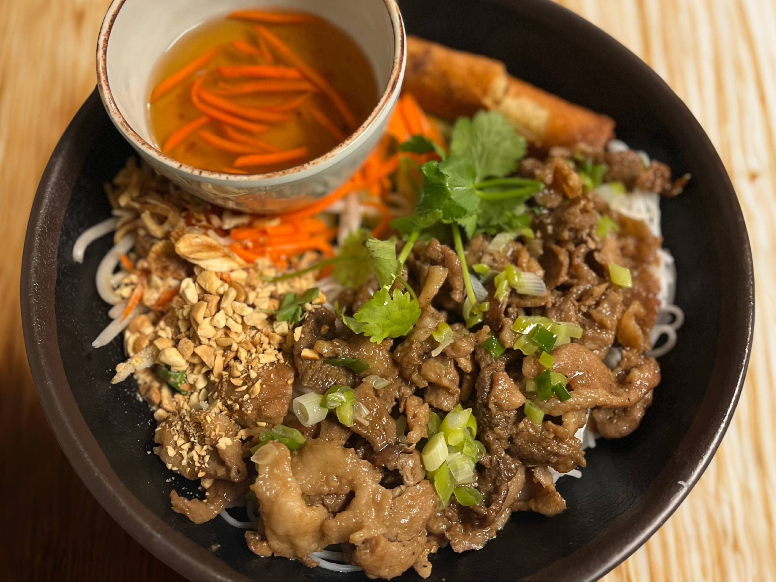 Bowl of Asian beef and noodle dish with chopped peanuts, shredded vegetables, fresh cilantro, and a small bowl of broth with carrots, served on a wooden table.