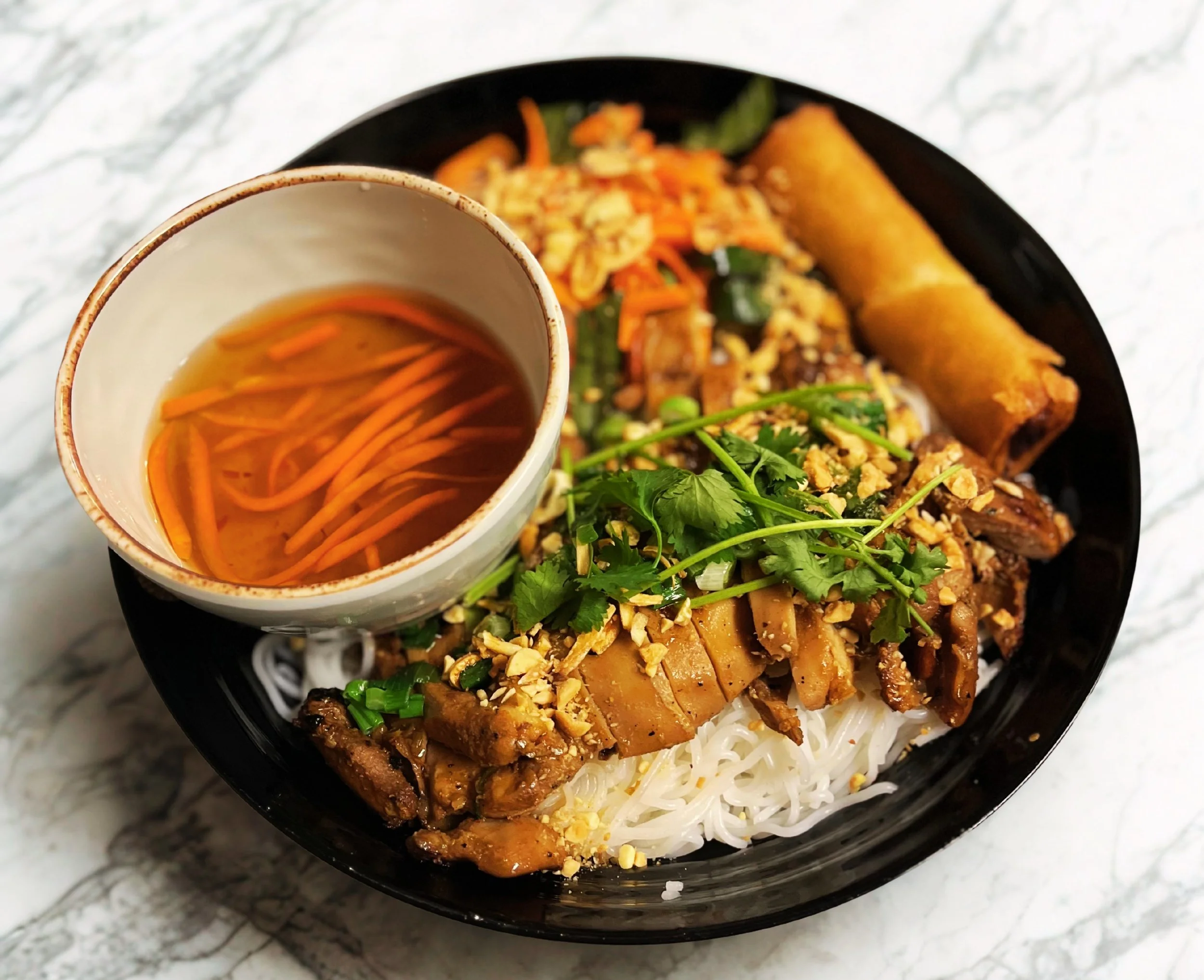 Bowl of Vietnamese pho with rice noodles, grilled meat, cilantro, chopped peanuts, and spring roll, accompanied by a side of tea with shredded carrots.