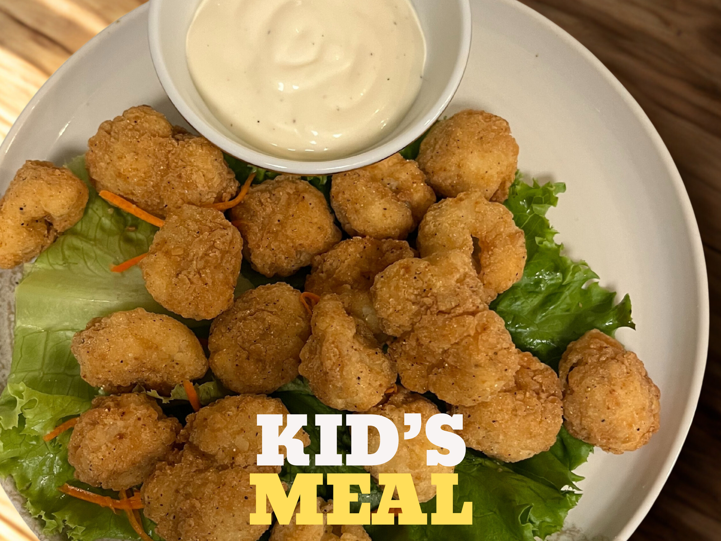 Bowl of ranch dressing, fried chicken nuggets, and leafy greens on a white plate with a wooden table in the background. Kid's meal text overlay.