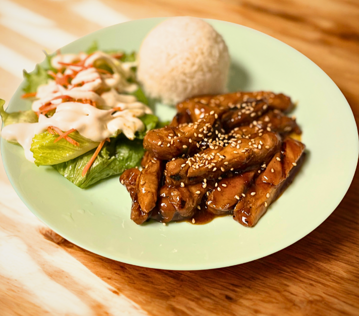 Chicken stir-fry with sesame seeds, white rice ball, and salad with lettuce and shredded vegetables on a green plate.
