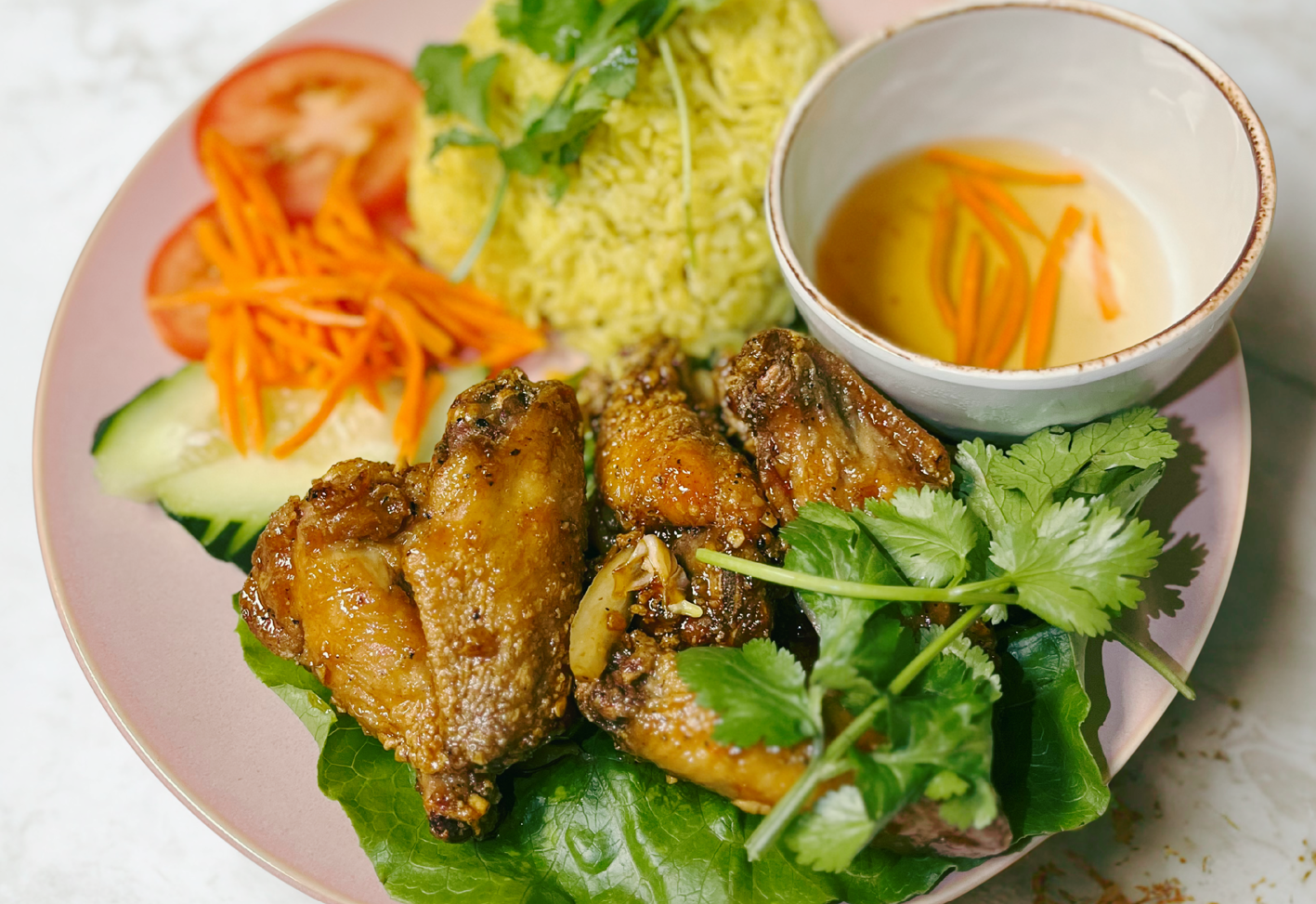 Plate with fried chicken wings, sliced tomatoes, cucumbers, shredded carrots, mashed potatoes with herbs, and a small bowl of dipping sauce with carrots, accompanied by fresh cilantro and greens.