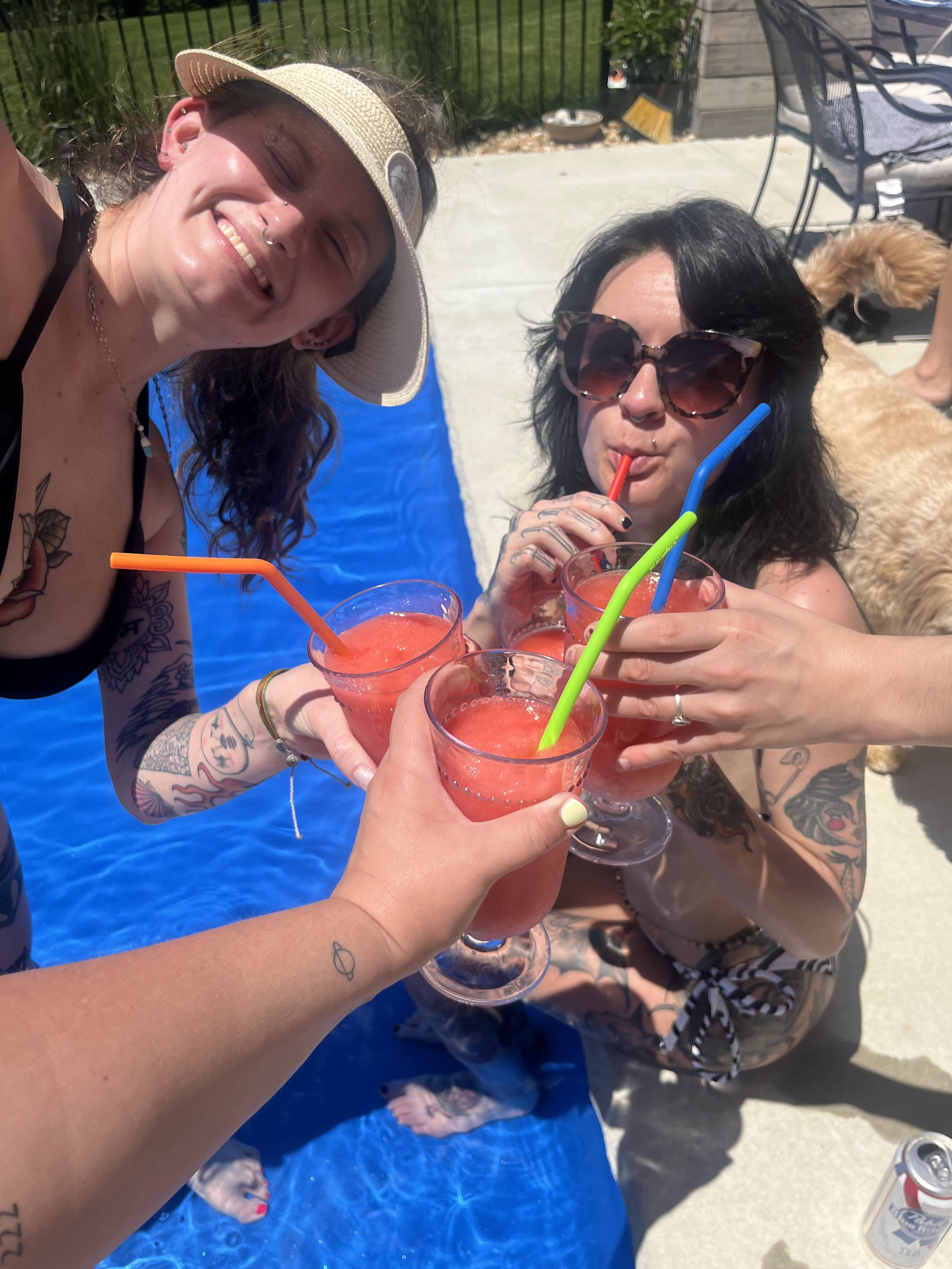 Four people clinking glasses of pink watermelon drinks with colorful straws at a poolside gathering on a sunny day.