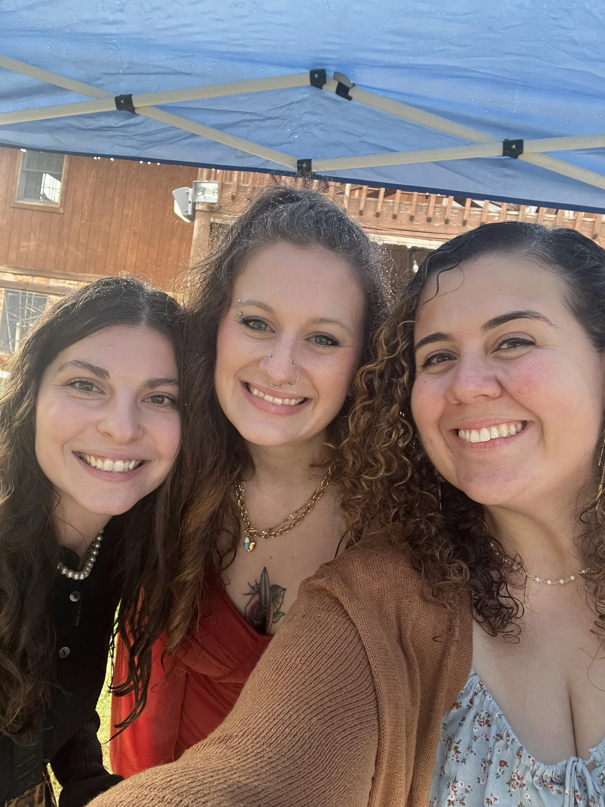 Three women smiling for a selfie outdoors under a blue canopy, with a wooden house in the background.